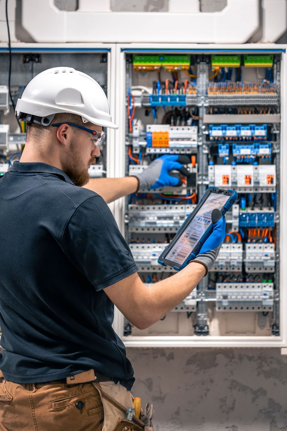 A man is working on an electrical box while using a tablet.