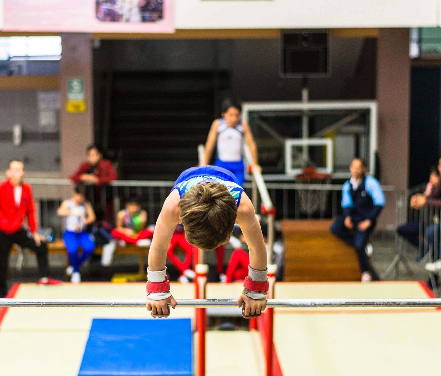 Boy in blue leotard performing gymnastics on horizontal bar in an indoor gym setting.