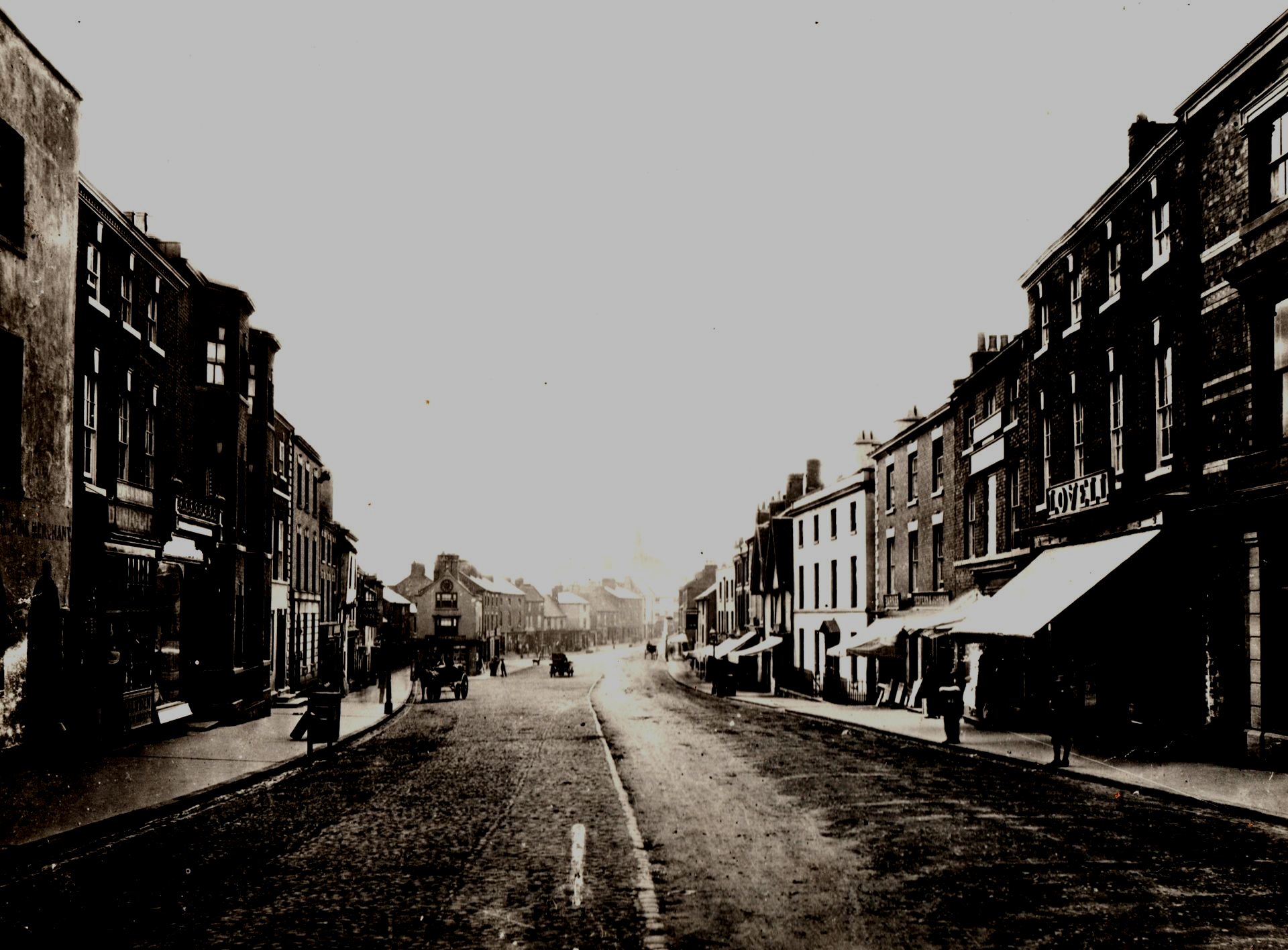Cobblestone street lined with buildings, horse-drawn carriages, and pedestrians in a vintage photo.