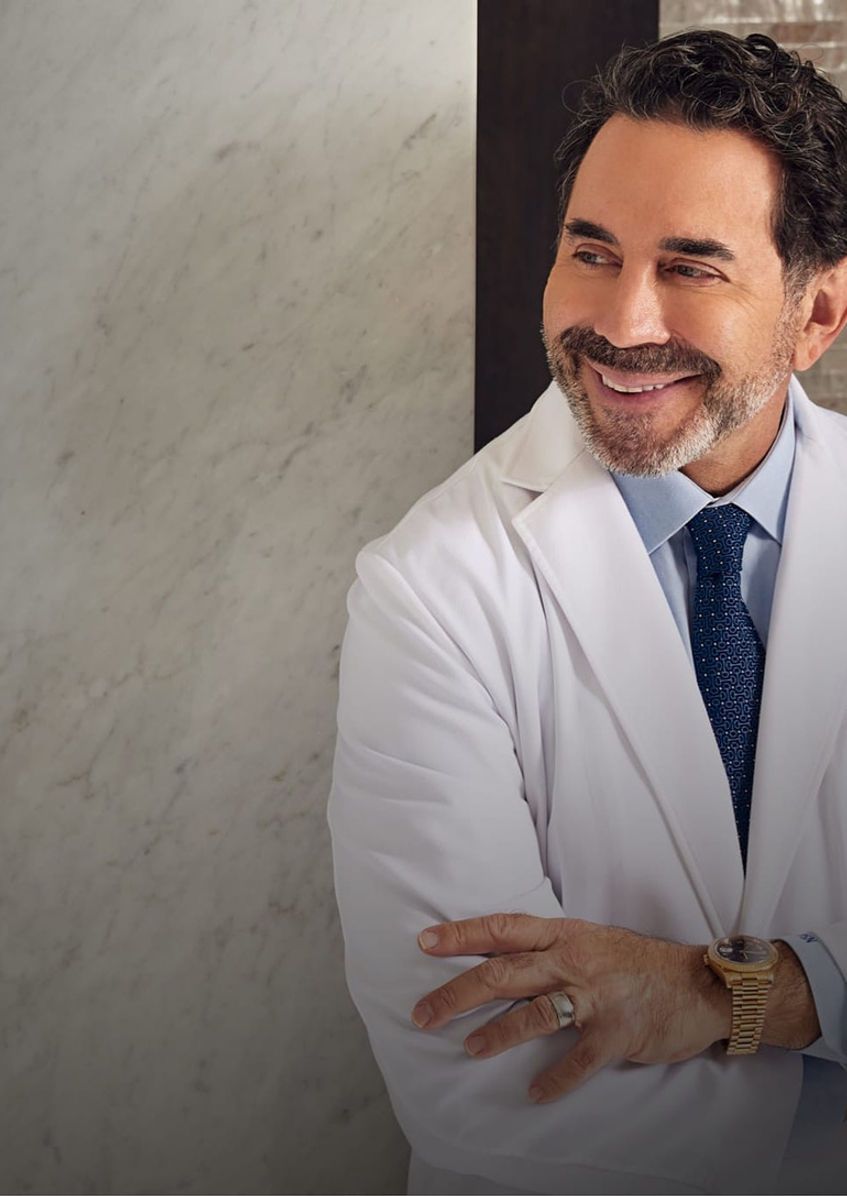 A man in a white coat and tie is smiling while standing with his arms crossed.