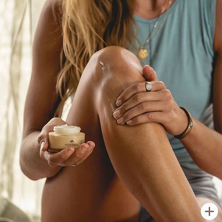 A woman is applying cream to her leg while sitting on a bed.