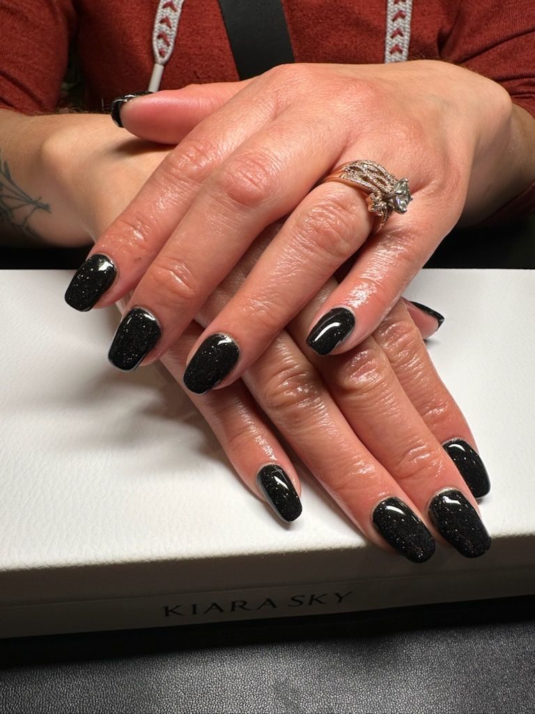 Hands with black, sparkly nail polish resting on a white surface, wearing a ring.