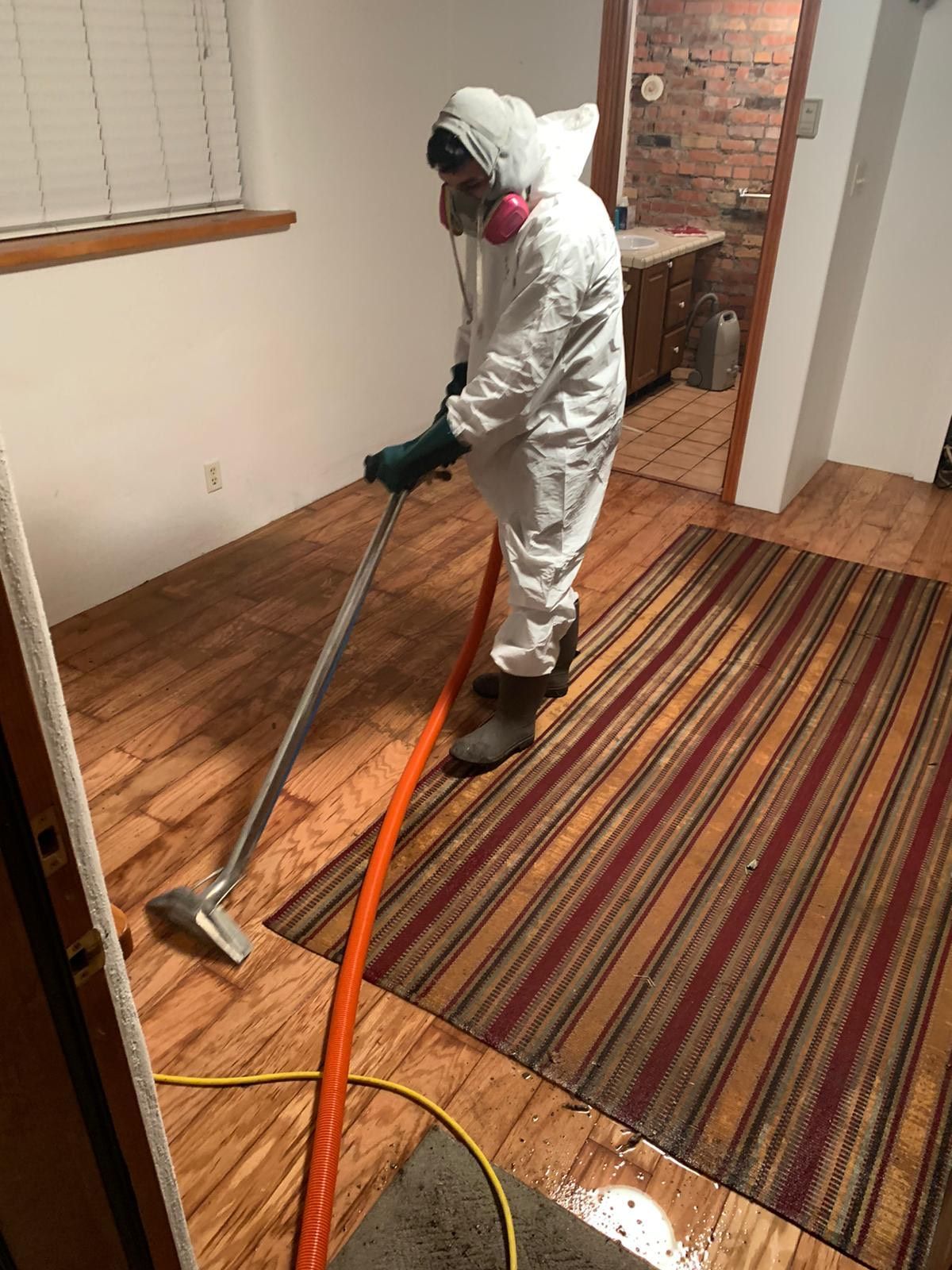 A man in a protective suit is cleaning a wooden floor with a vacuum cleaner.