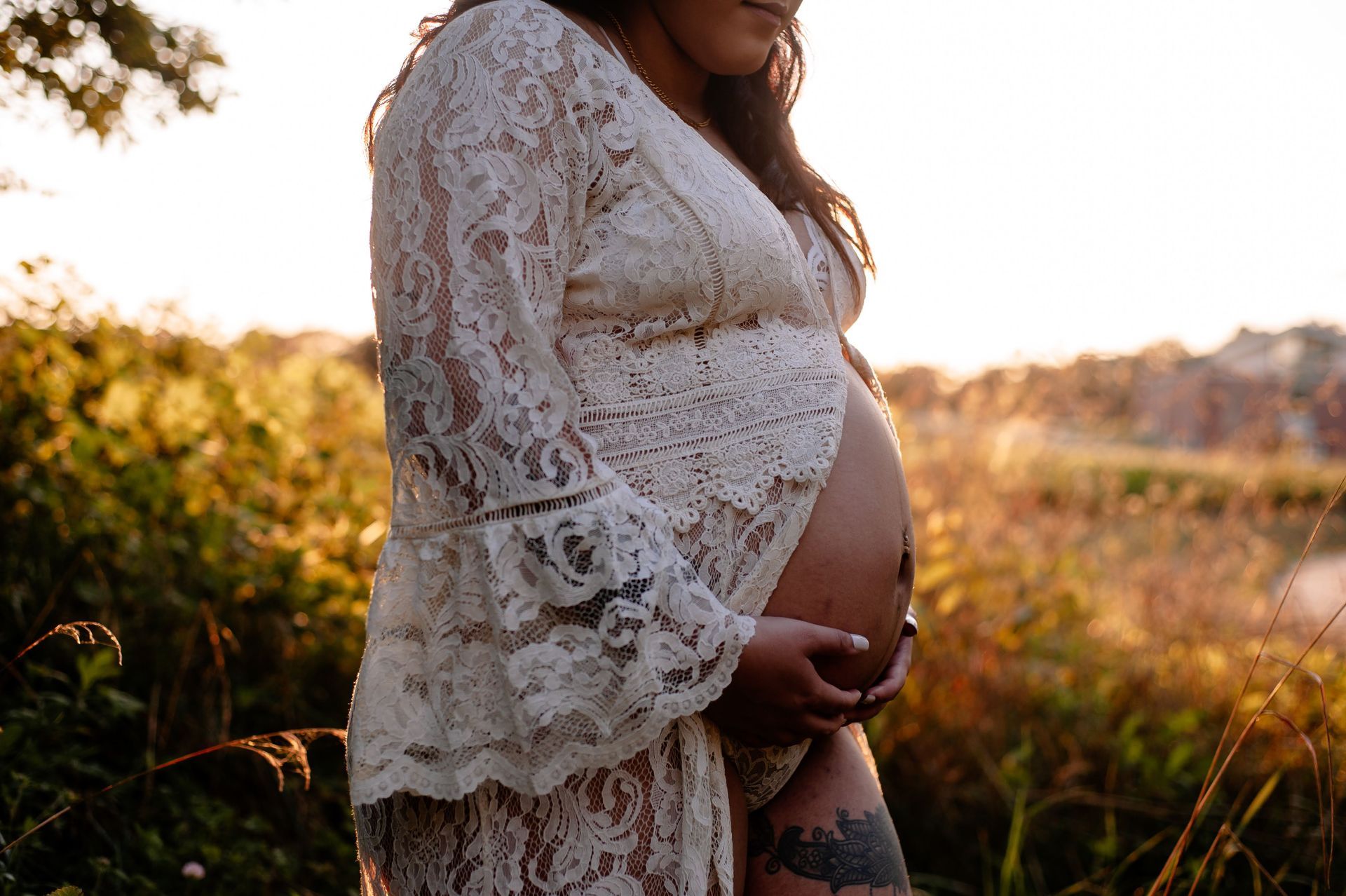 A pregnant woman in a white lace dress is holding her belly in a field.