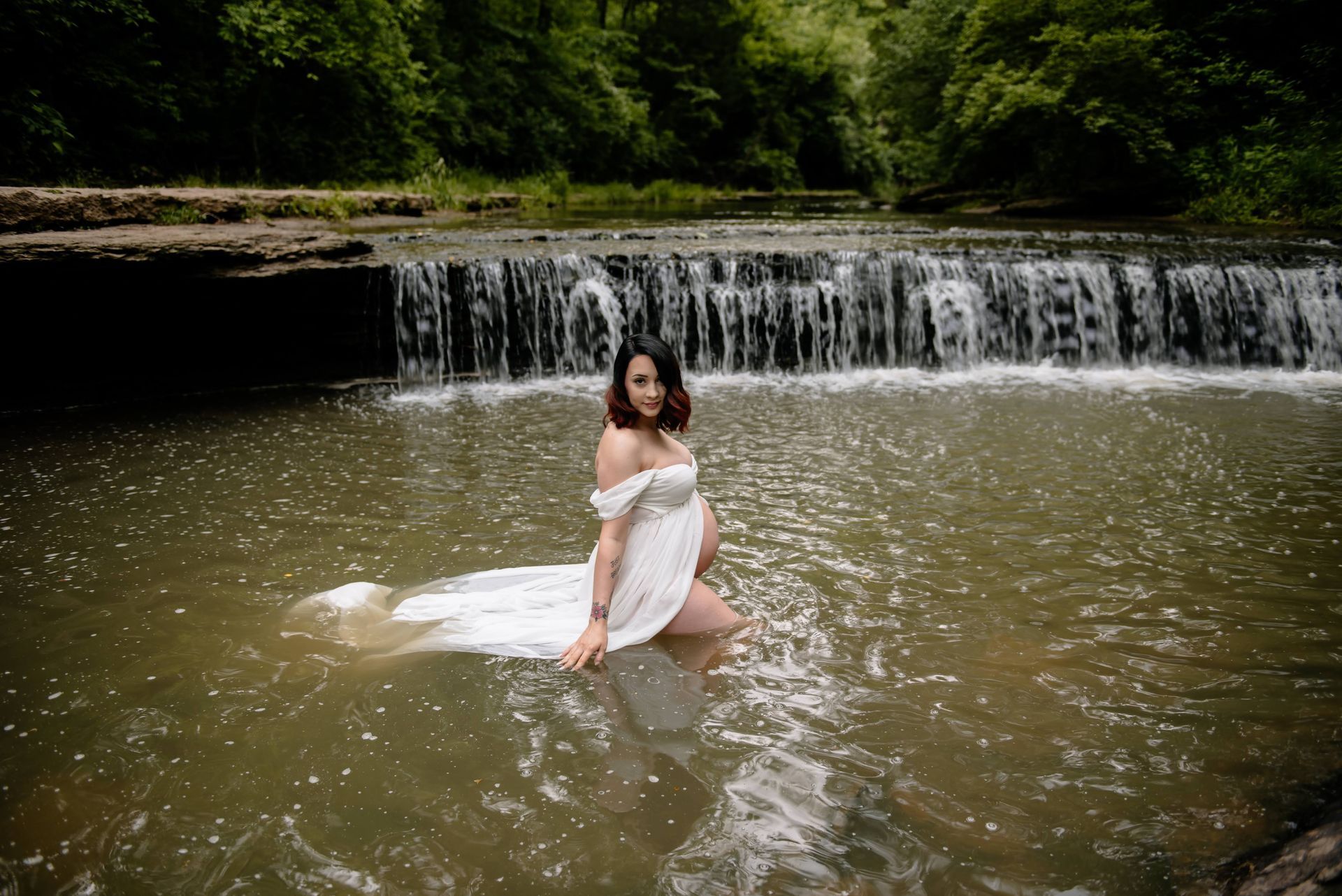A pregnant woman in a white dress is standing in the water near a waterfall.