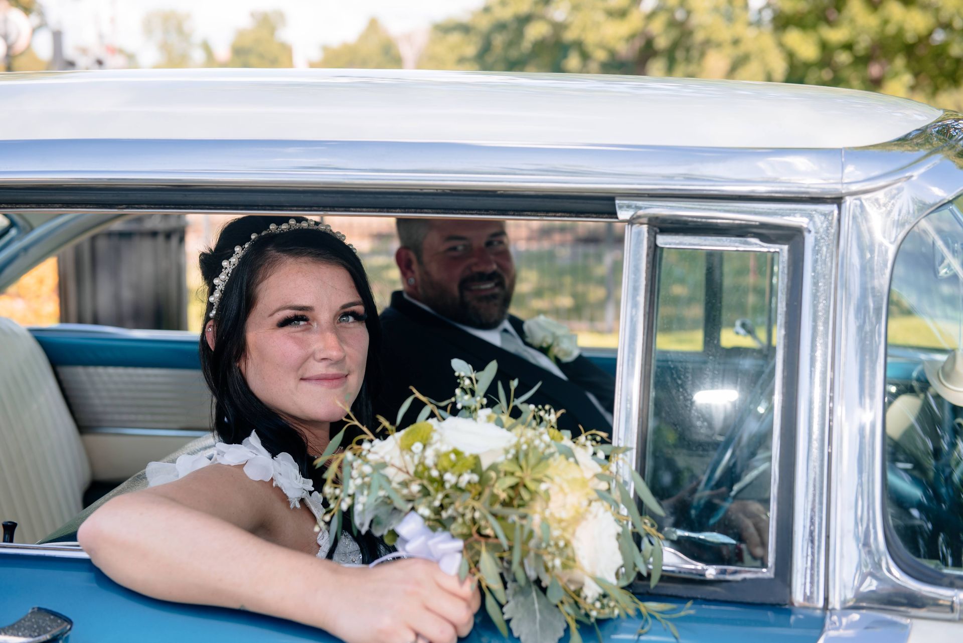 A bride and groom are sitting in a blue car . the bride is holding a bouquet of flowers.