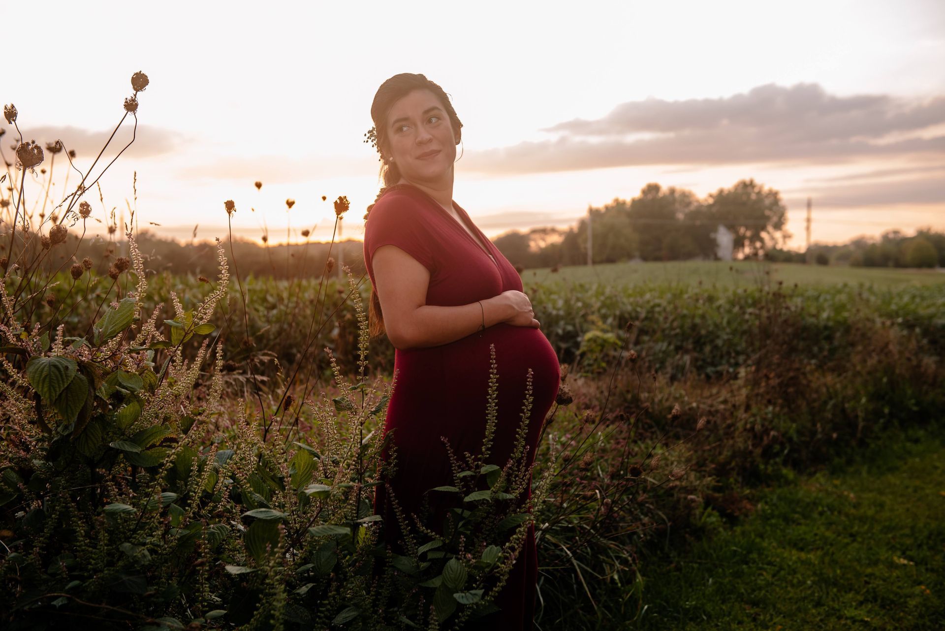 A pregnant woman is standing in a field at sunset holding her belly.