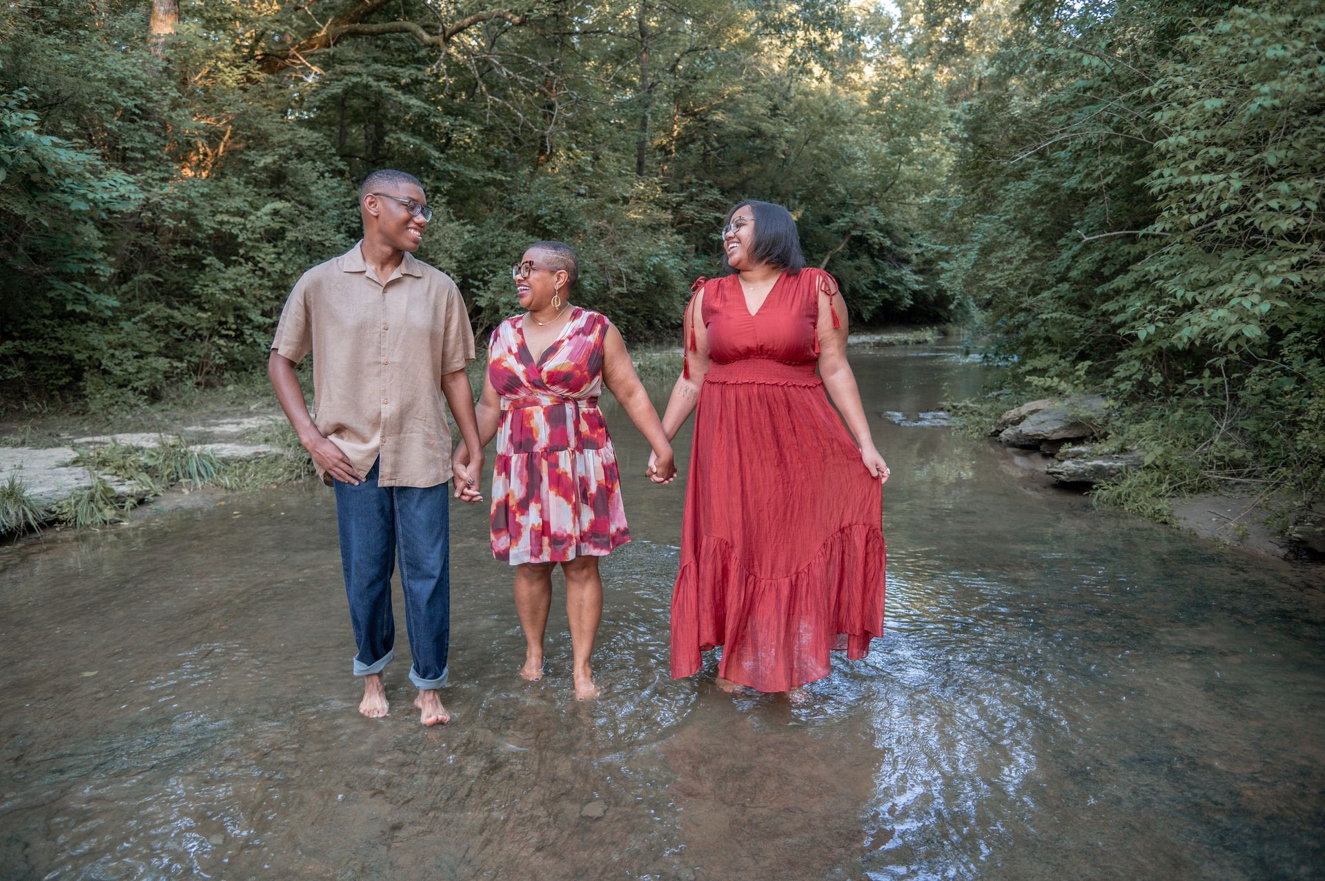 A man and two women are walking through a stream holding hands.