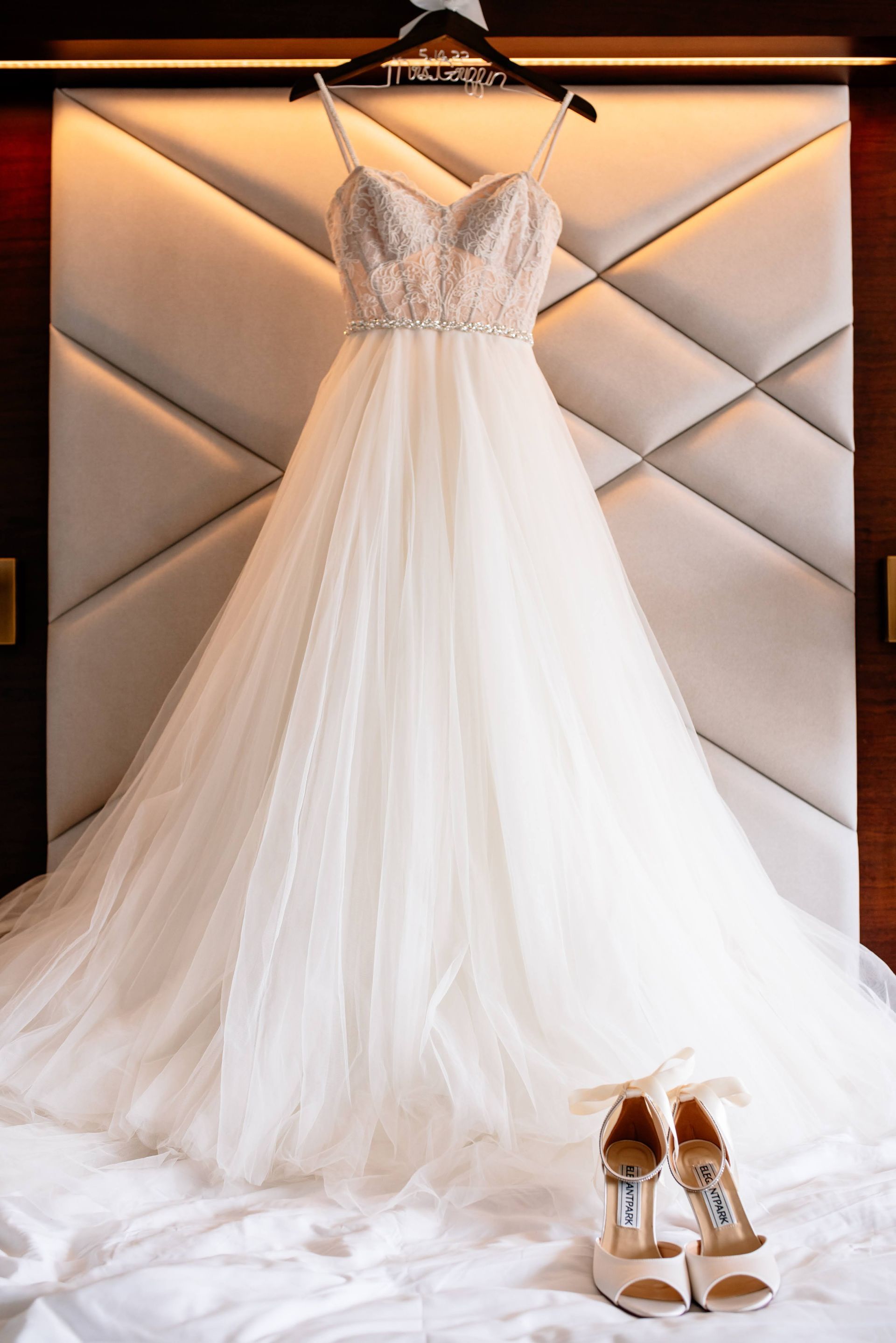 A wedding dress is hanging on a hanger on a bed next to a pair of shoes.