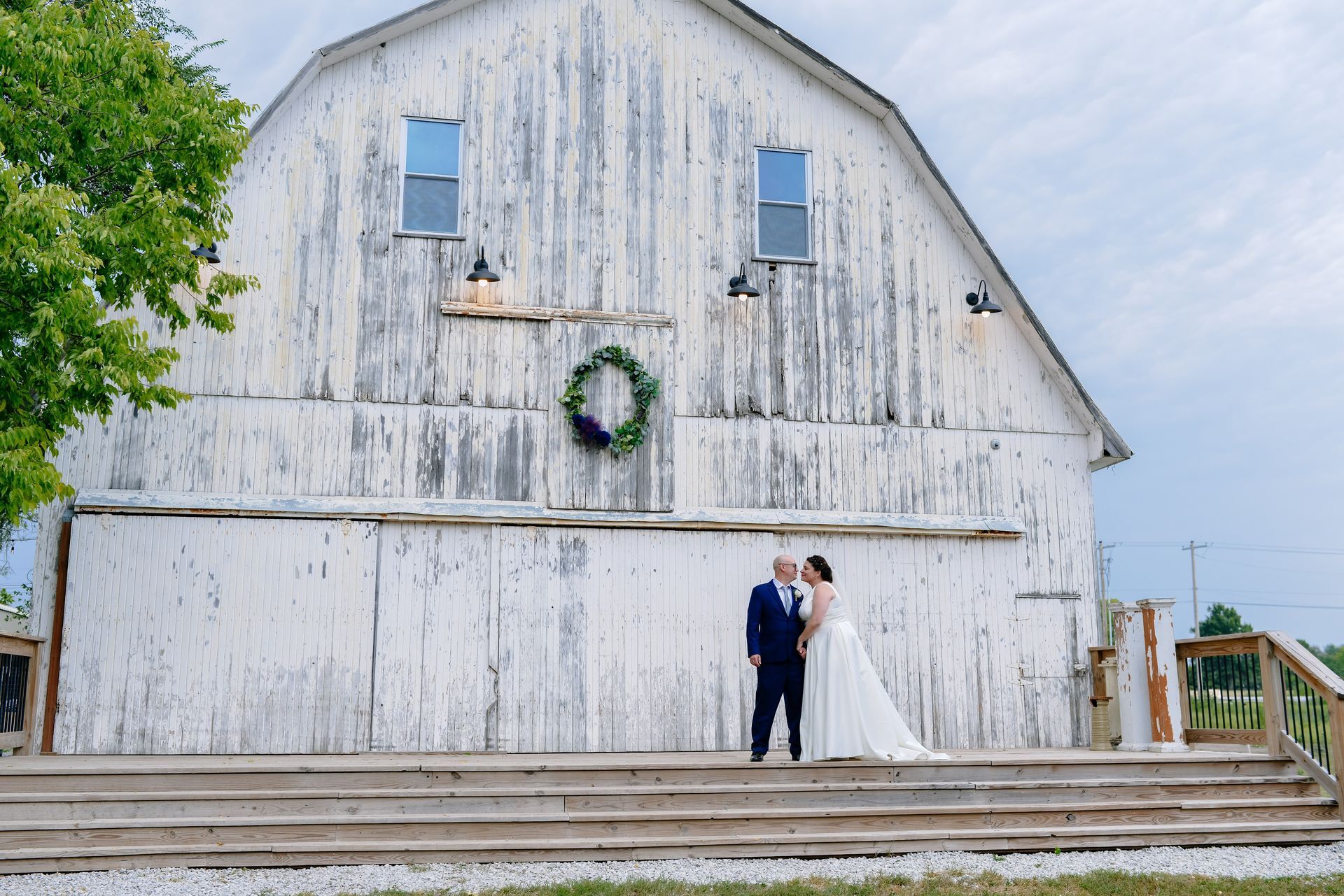 A bride and groom are kissing in front of a white barn.