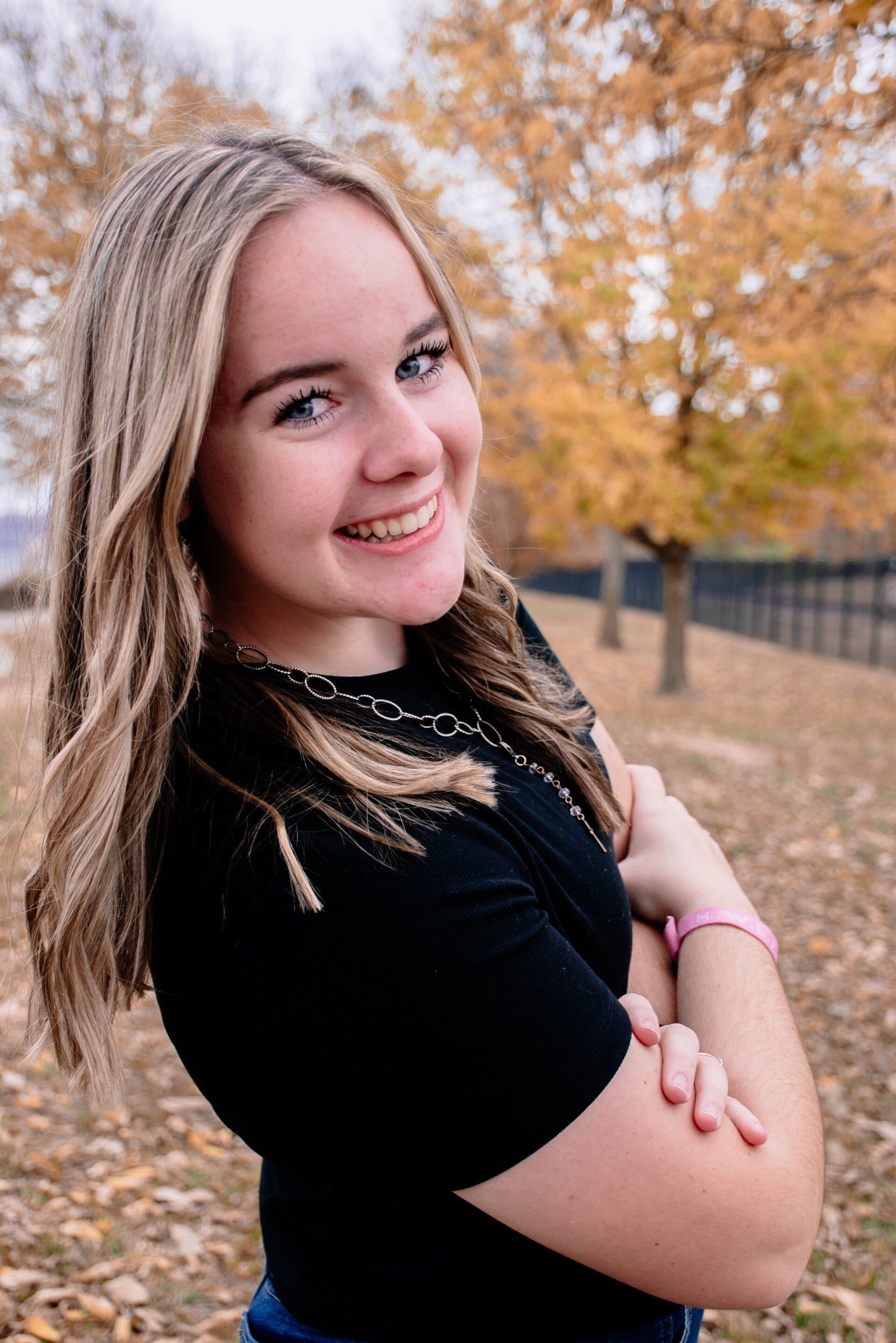 A young woman in a black shirt is standing in a park with her arms crossed and smiling.