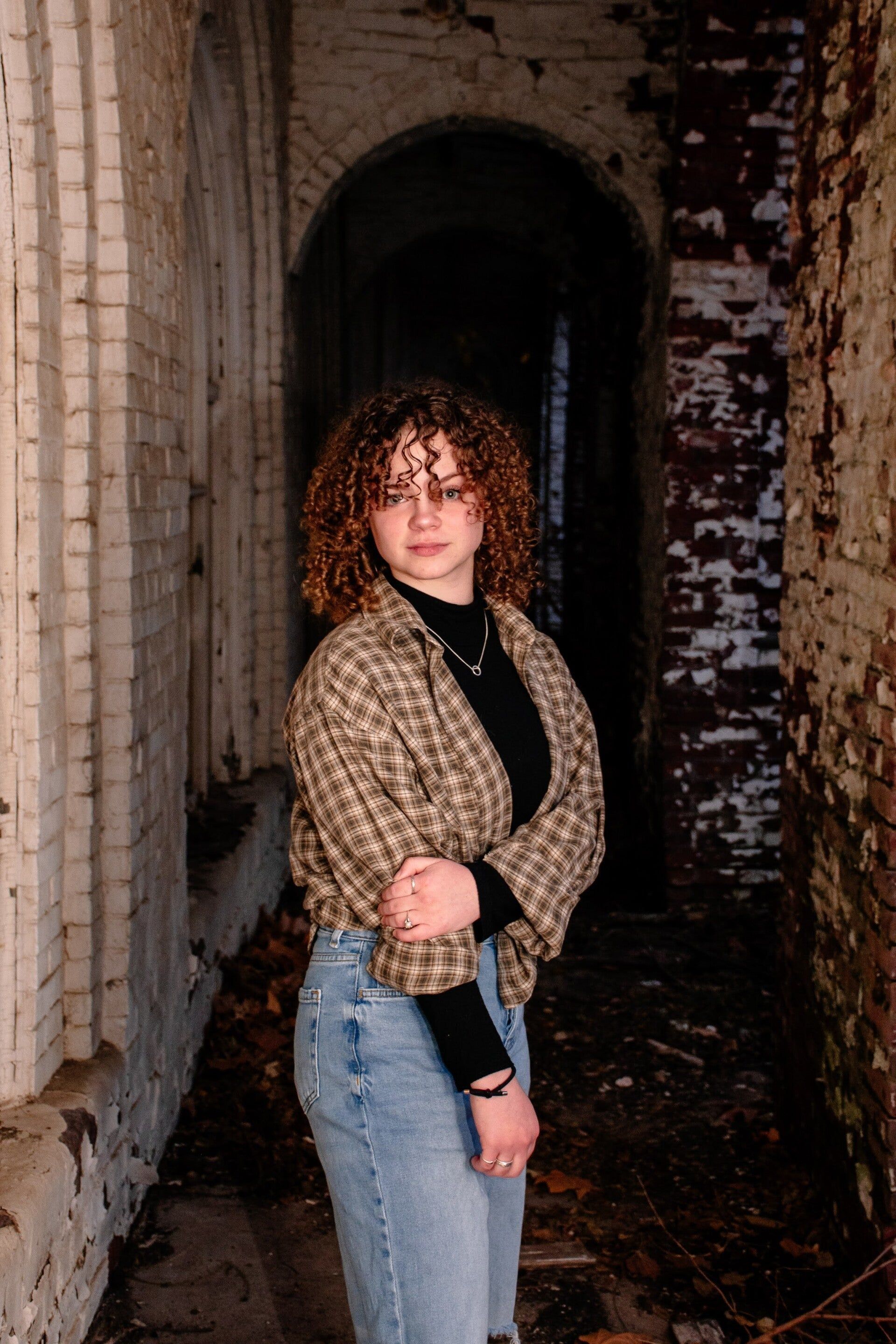 A woman with curly hair is standing in a dark hallway next to a brick wall.