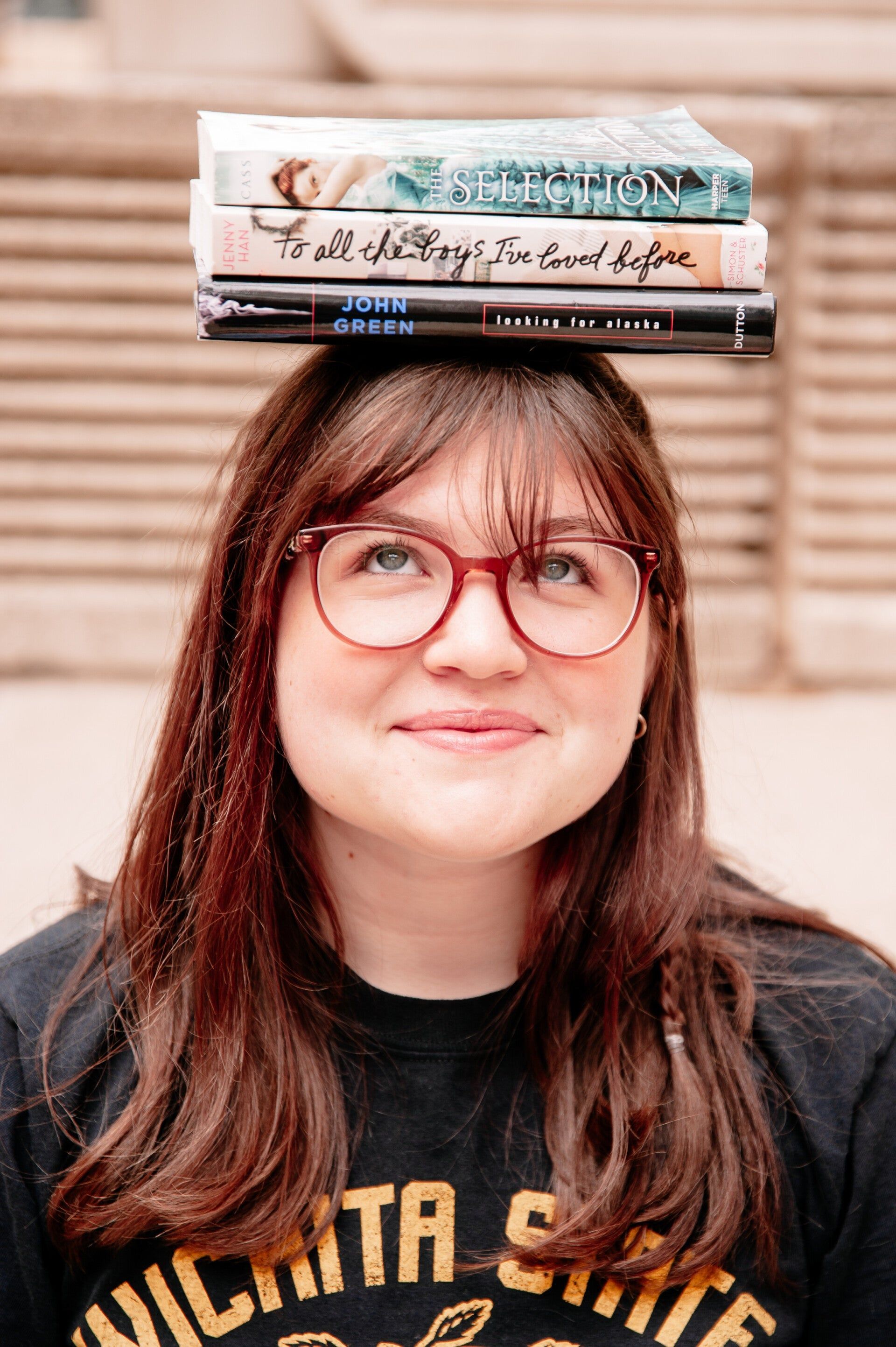 A woman with glasses is holding a stack of books on her head.