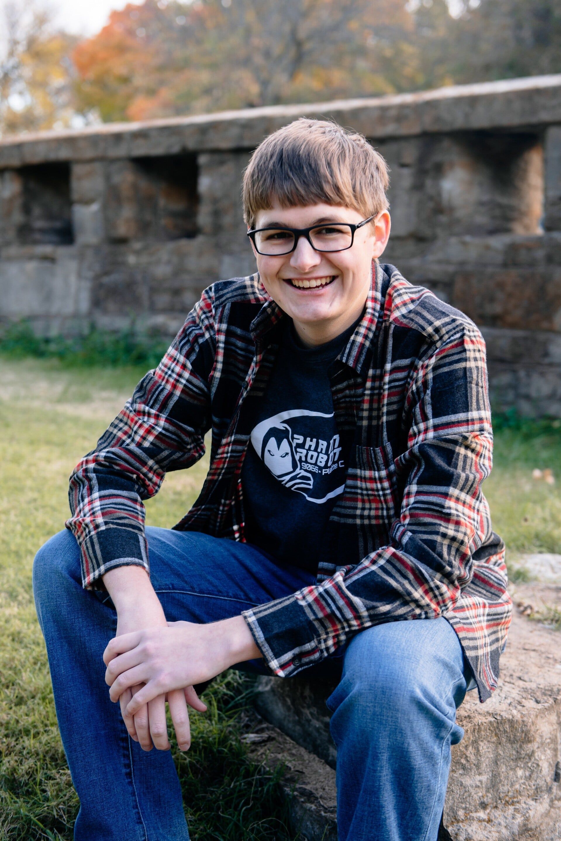 A young man wearing glasses and a plaid shirt is sitting on a rock.
