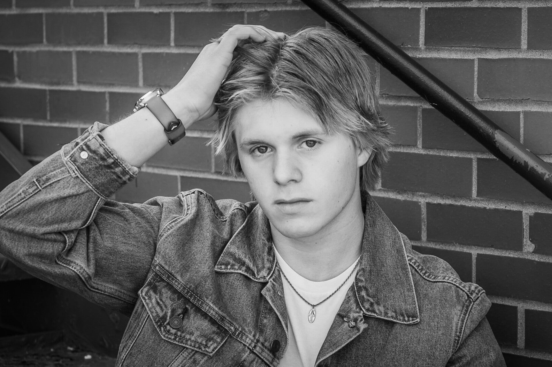 A young man in a denim jacket is sitting in front of a brick wall.