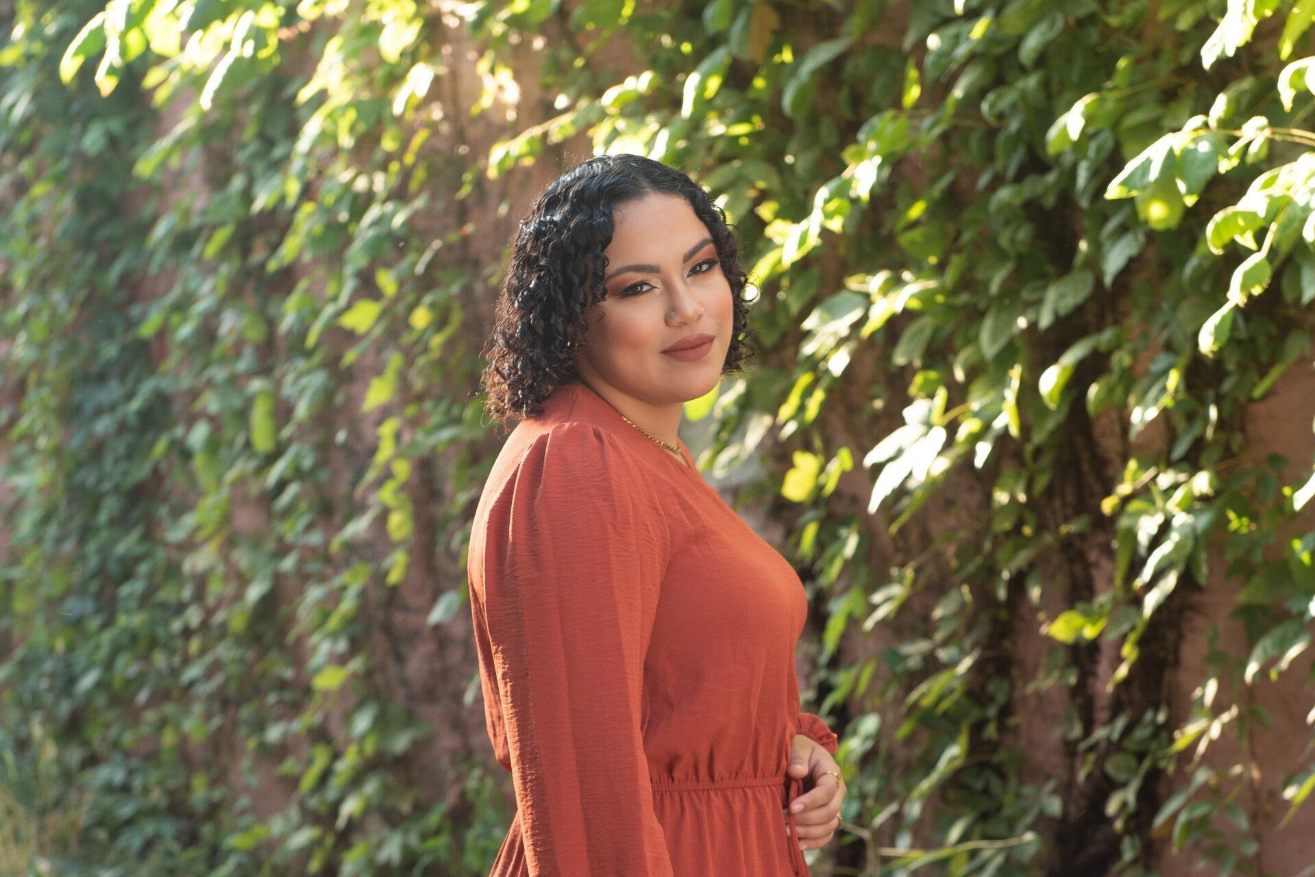 A woman in a red dress is standing in front of a ivy wall.