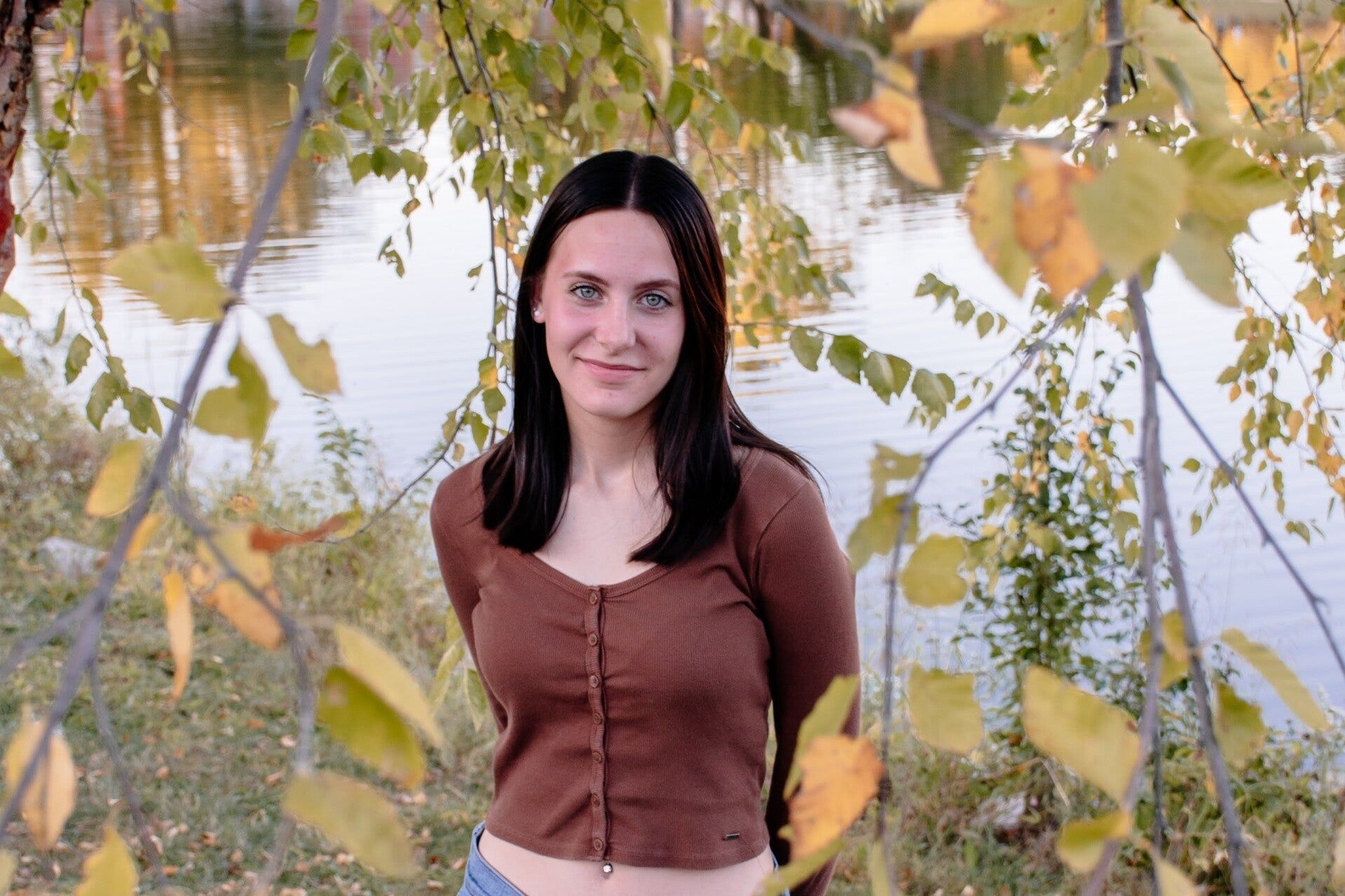 A woman in a brown sweater is standing in front of a body of water.