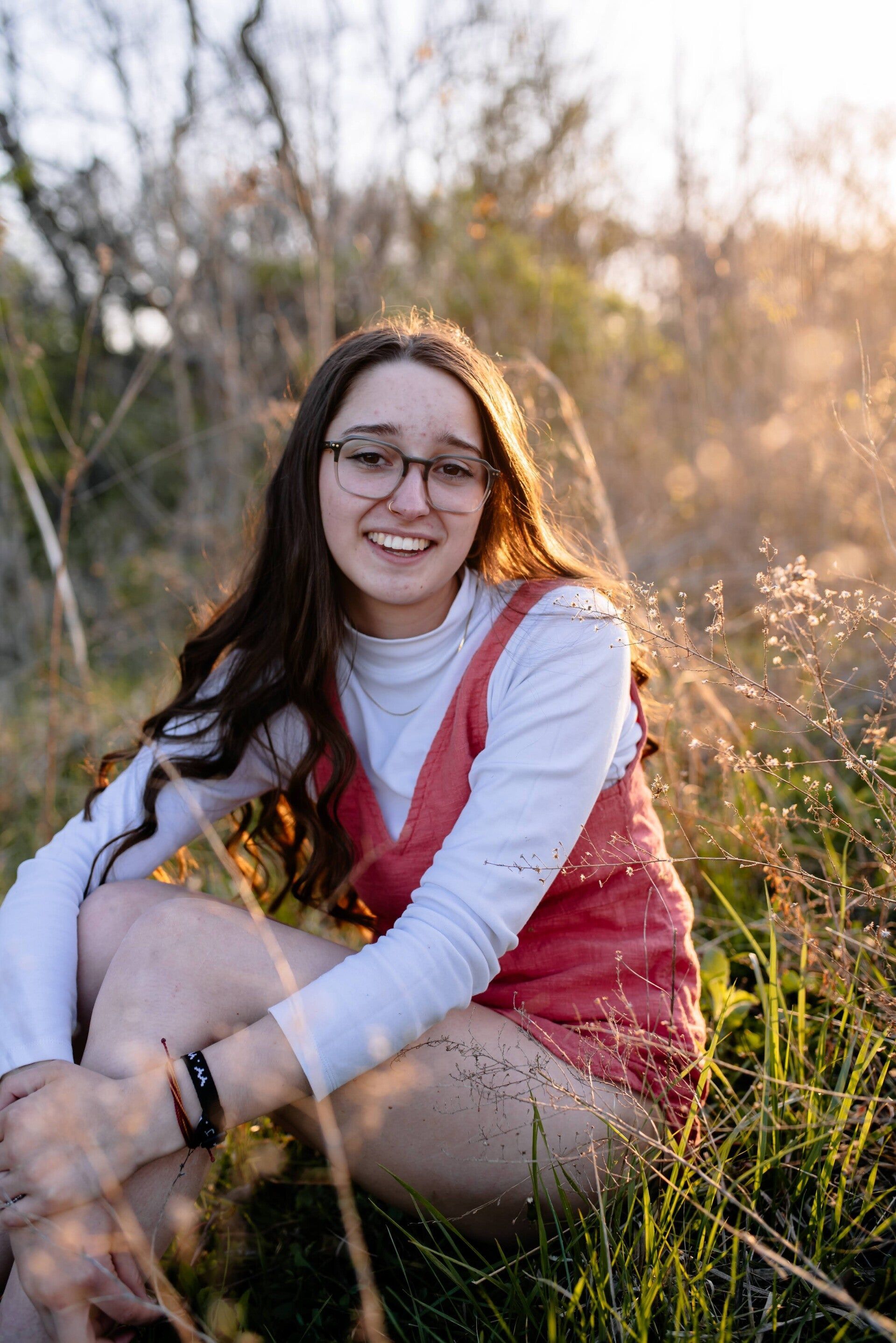 A young woman wearing glasses is sitting in a field of tall grass.