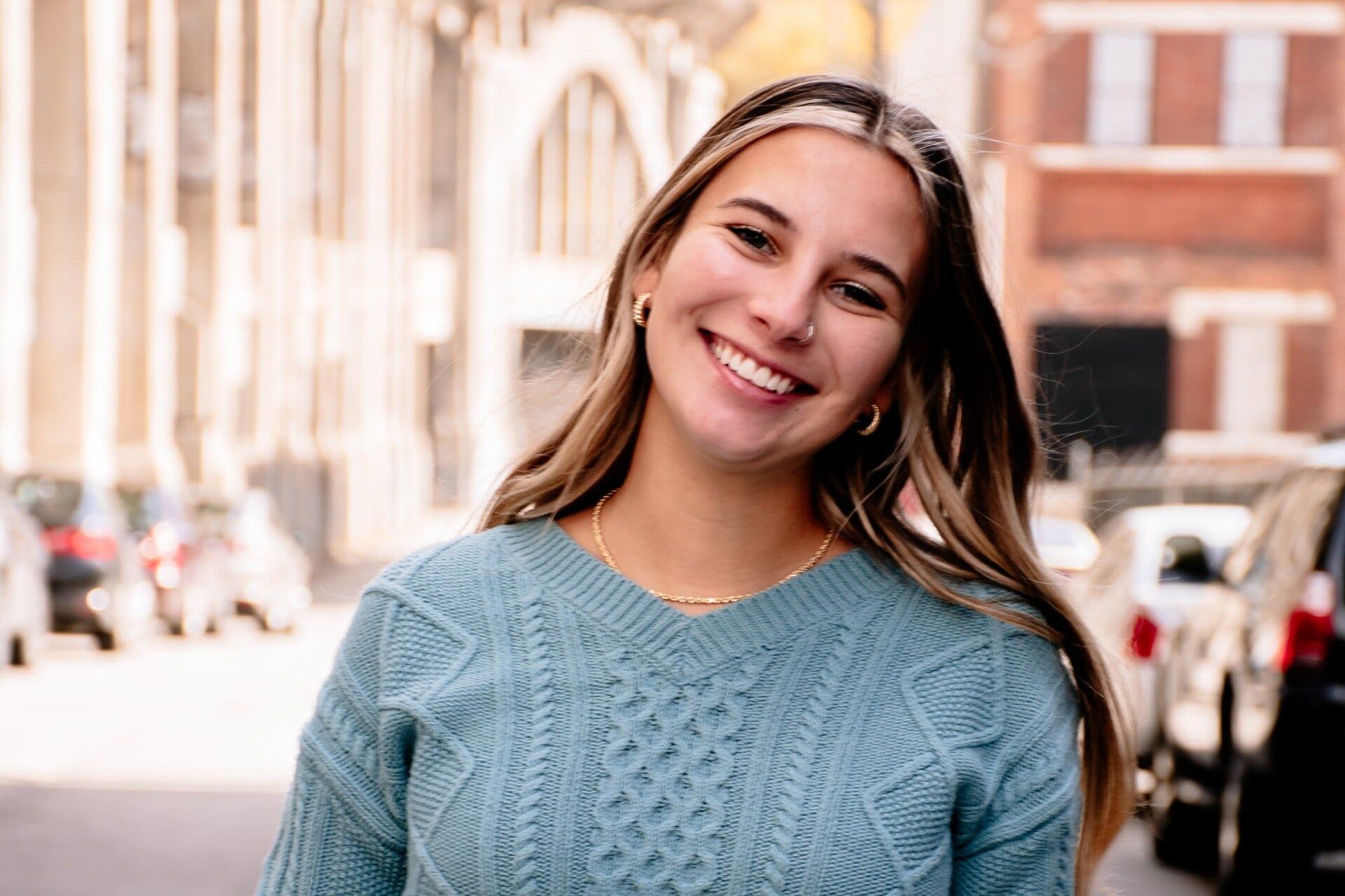 A woman in a blue sweater is smiling while standing on a city street.