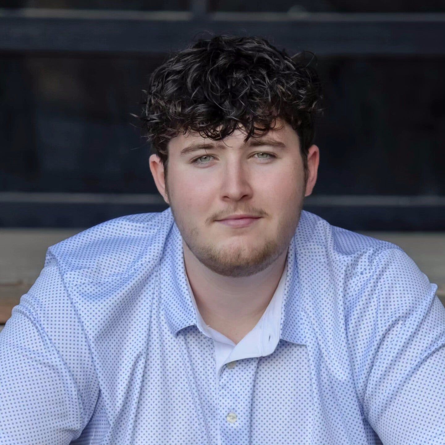 A young man with curly hair and a beard is wearing a blue shirt and looking at the camera.