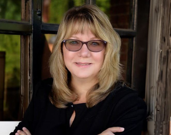 A woman wearing glasses and a black shirt is standing in front of a window.