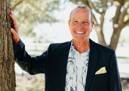 A man in a suit is leaning against a tree and smiling.