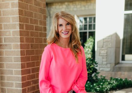 A woman in a pink shirt is standing in front of a brick building.