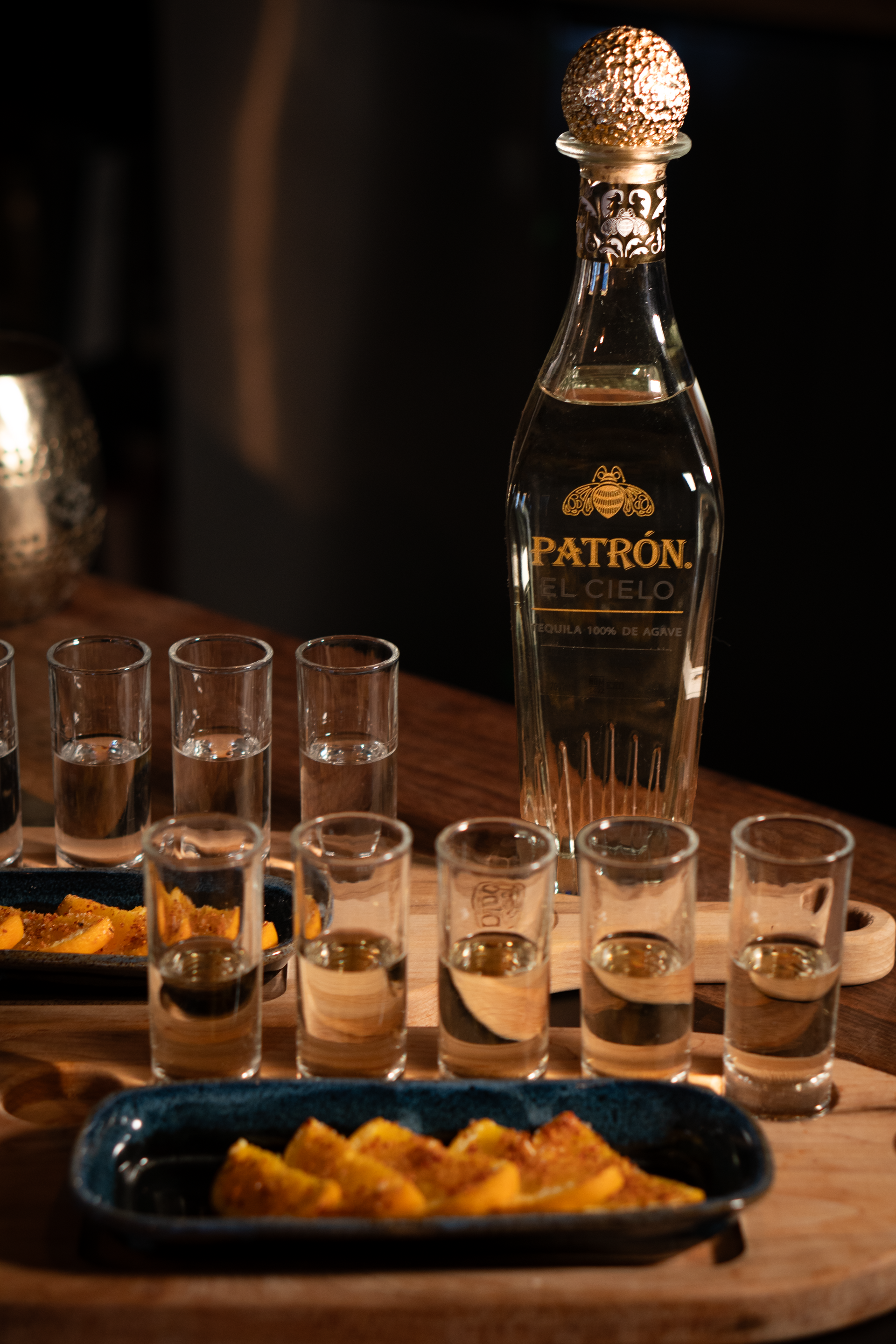 A bottle of patron tequila is sitting on a wooden table next to shot glasses and chips.