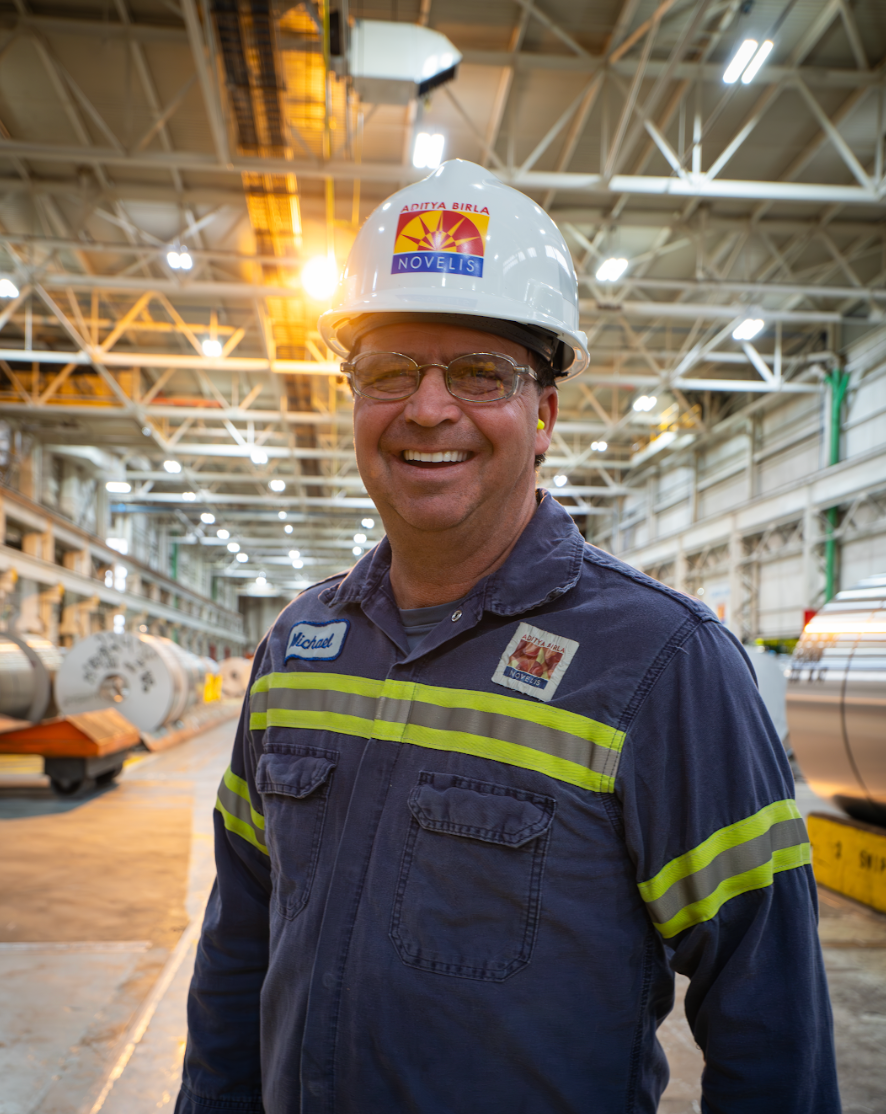 A man wearing a hard hat and overalls is smiling in a factory.