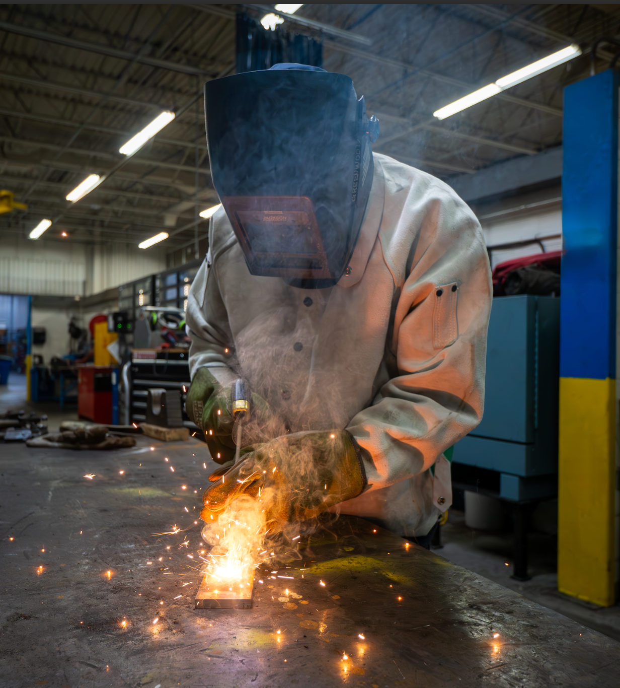 A man wearing a welding mask is welding a piece of metal in a factory.