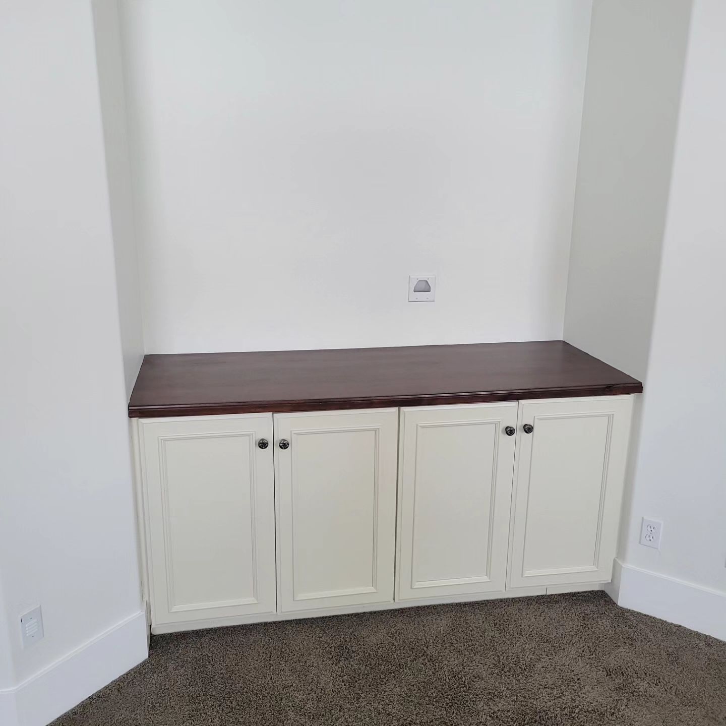 Built-in cabinet with four doors, topped with a dark surface. The cabinet is white, set in a nook.