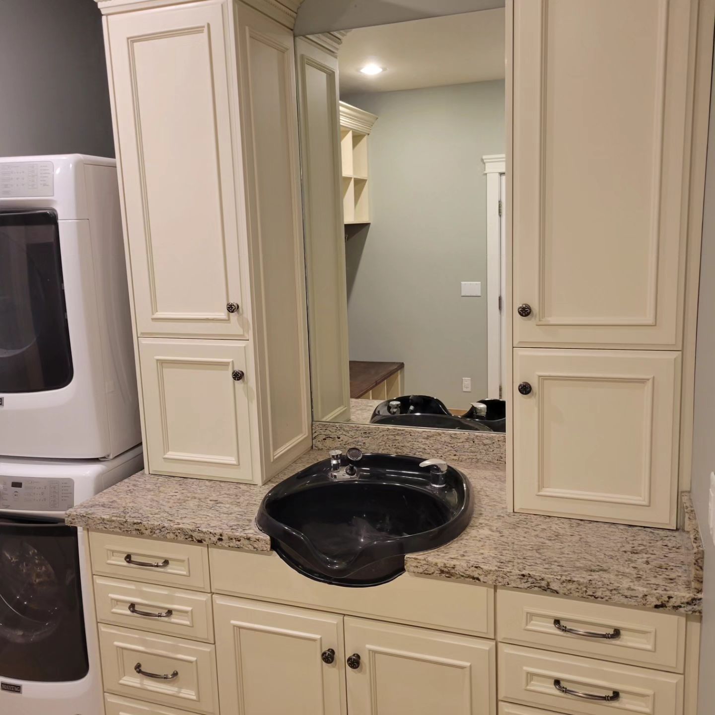 Salon station with black shampoo bowl, tan cabinets, granite countertop, and mirror. A washer/dryer are to the left.