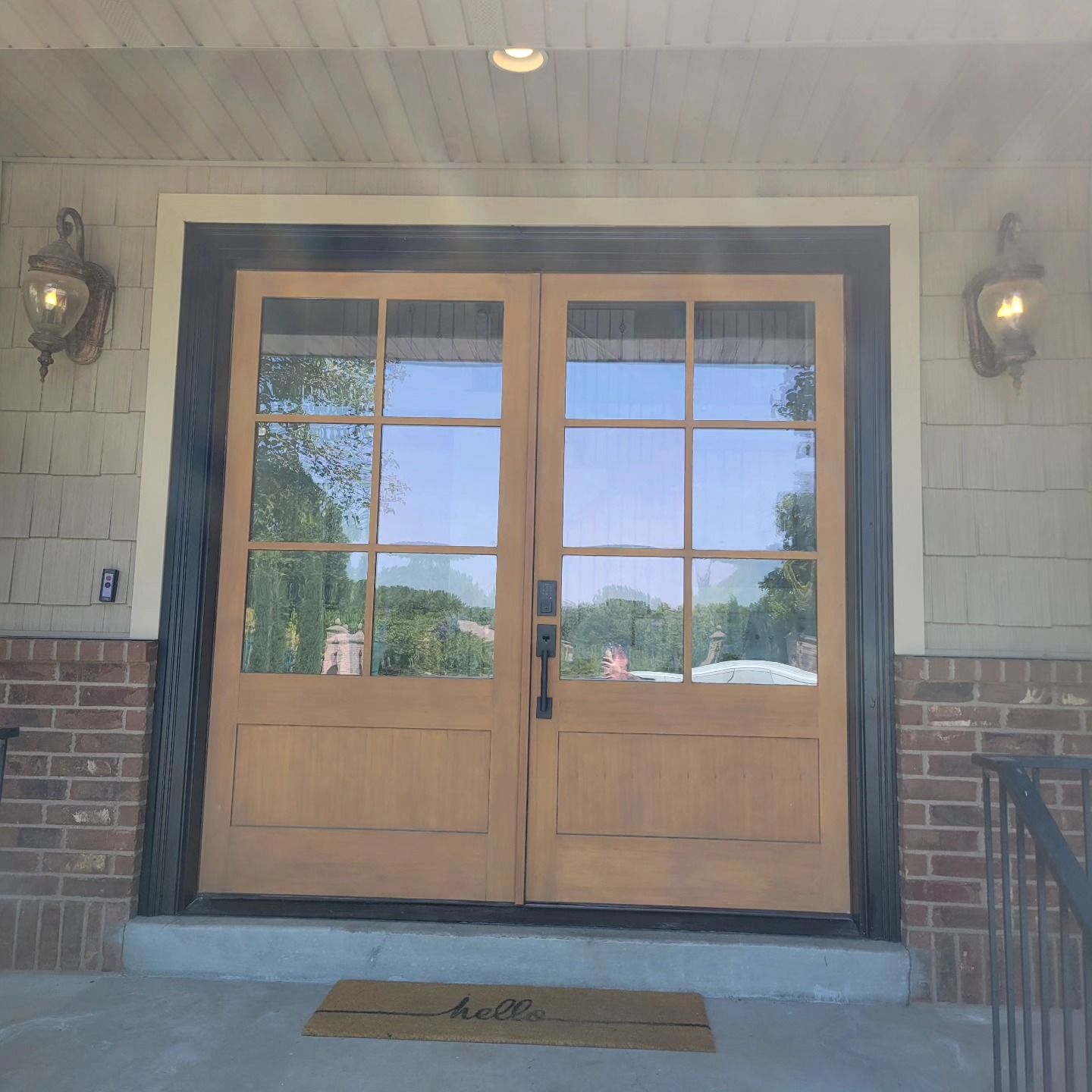 Double wooden front doors with glass panes, framed in black, on a brick and light-colored siding porch, with sconces.