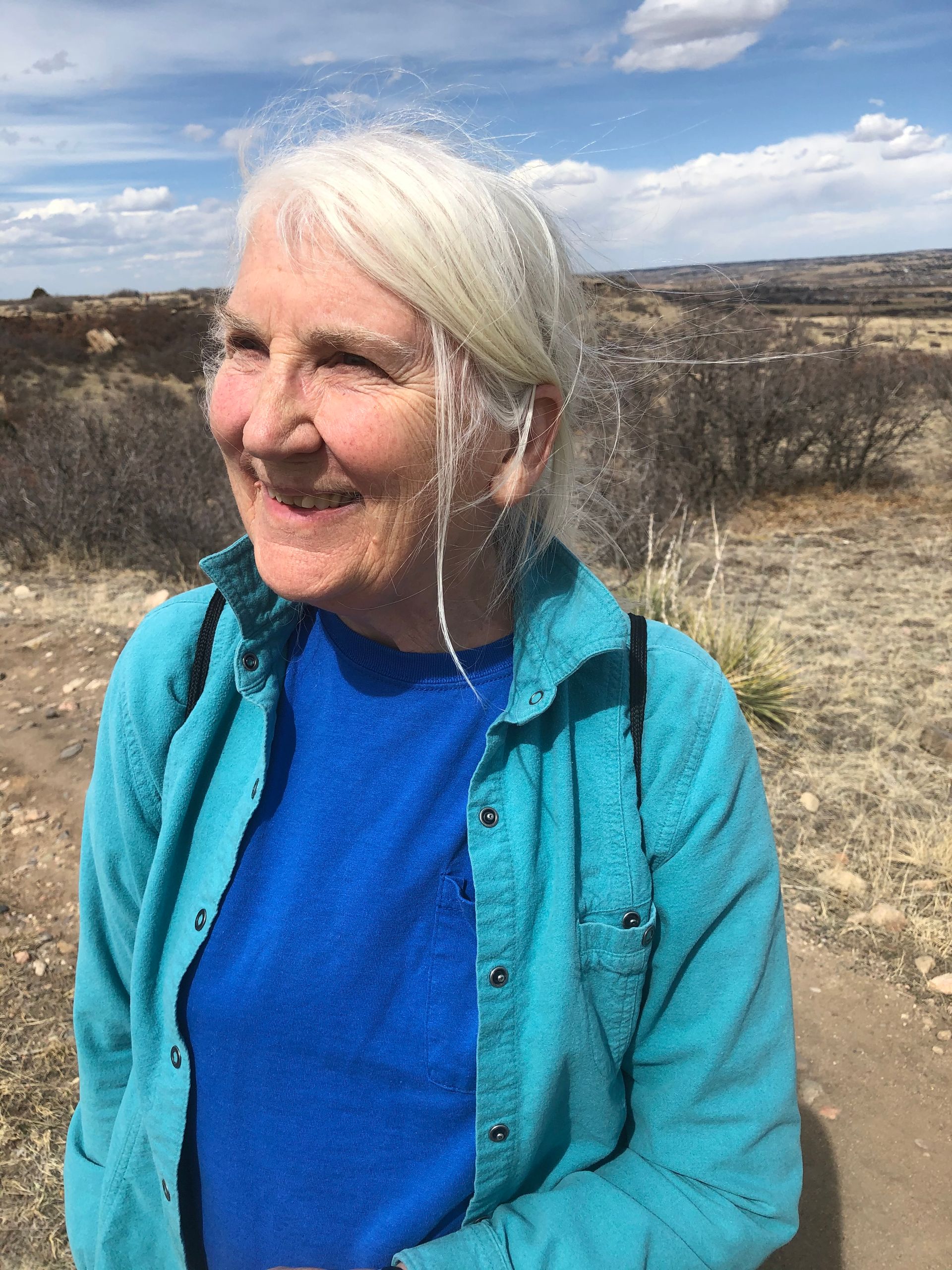 A woman in a blue shirt is smiling while standing on a dirt road.