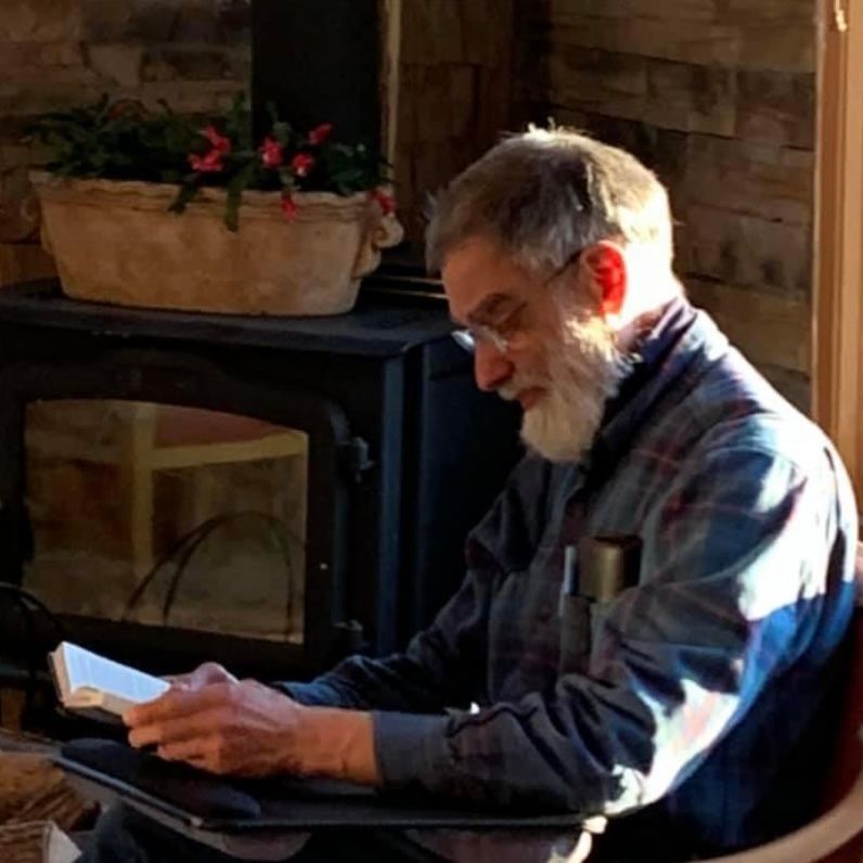 A man with a beard is reading a book in front of a fireplace