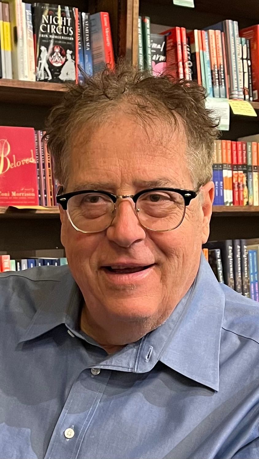 A man wearing glasses and a blue shirt is standing in front of a bookshelf.
