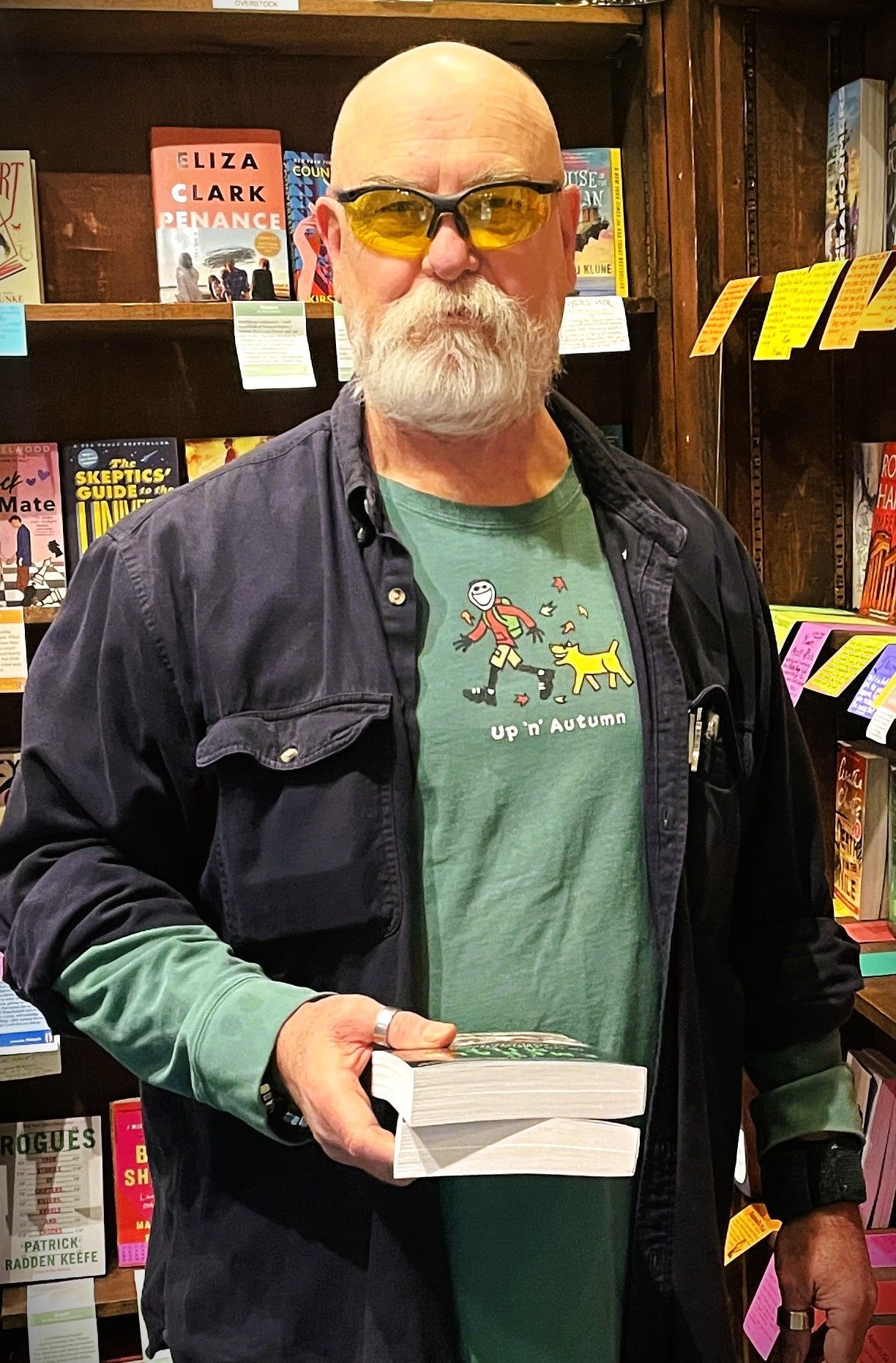 A bald man with a beard is holding two books in front of a bookshelf.
