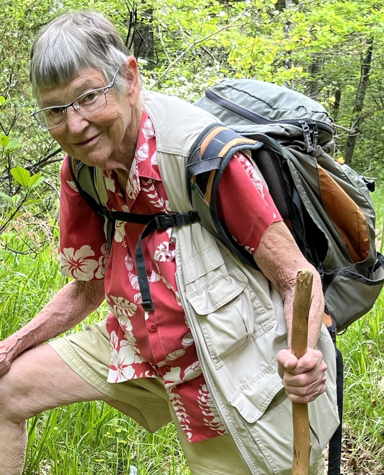 A man with a backpack and a cane is kneeling down in the woods.