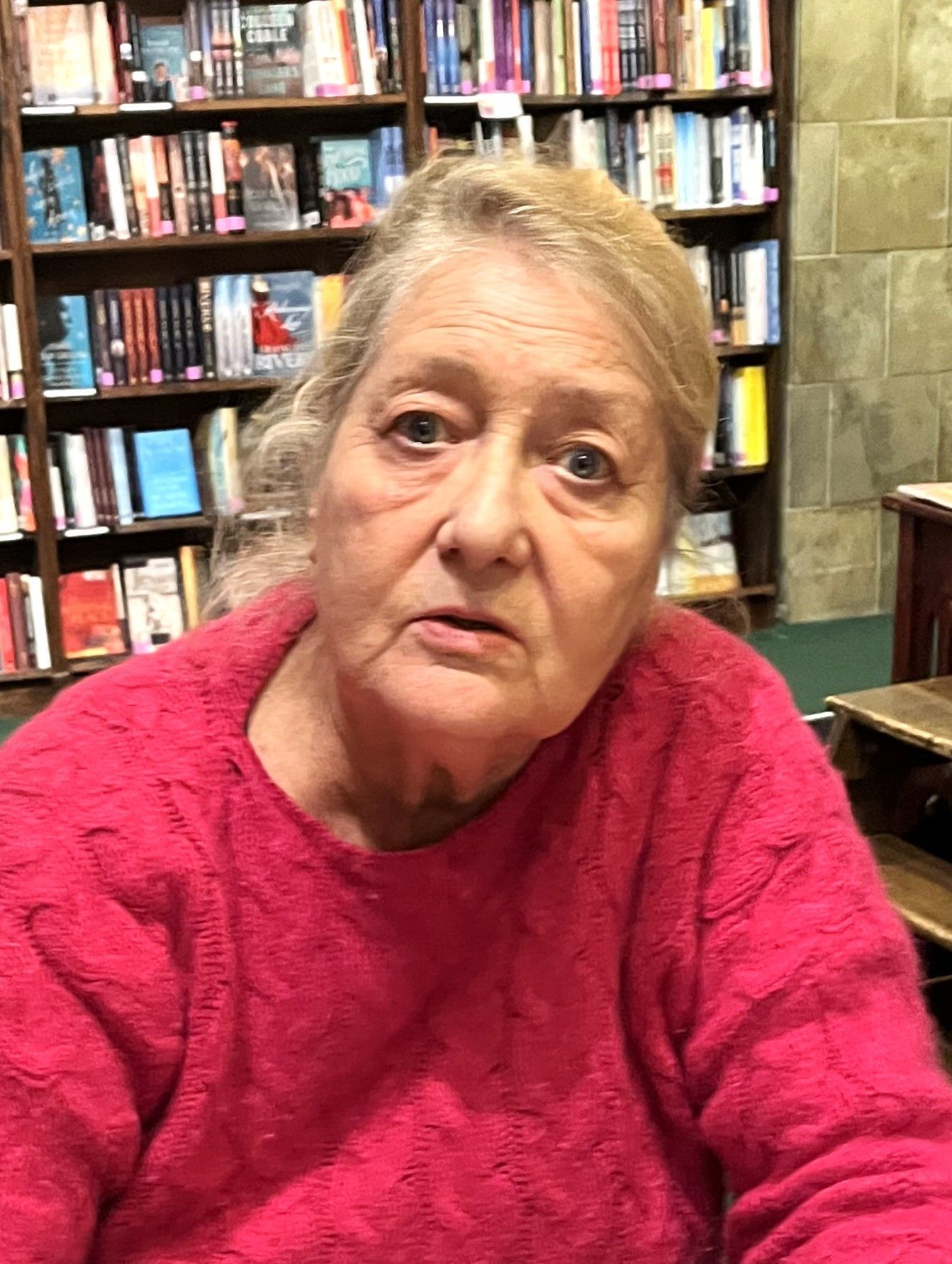 A woman in a pink sweater is sitting in front of a bookshelf.