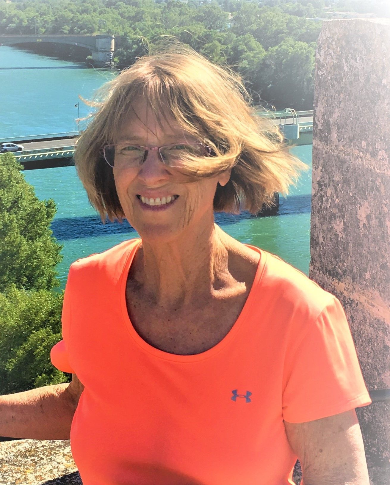 A woman wearing glasses and an orange shirt is smiling in front of a body of water.
