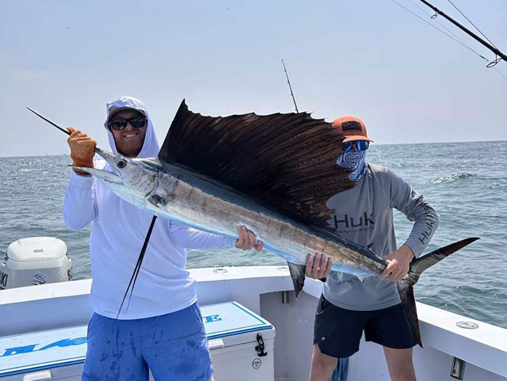 Two men in baseball caps and sunglasses standing on a deep sea fishing charter out of Hilton Head and holding a sizeable Sailfish between them on a clear day