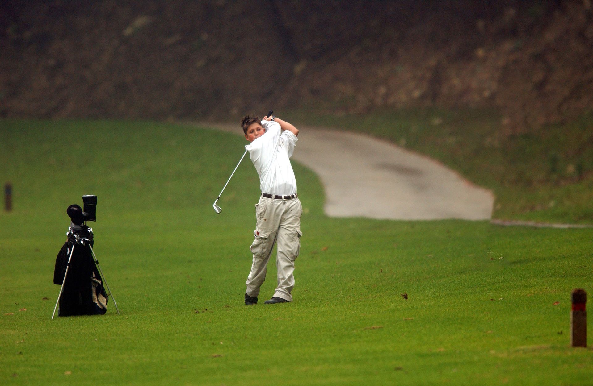 Golfer mid-swing on a green fairway, with a golf bag beside him and a winding path in the background