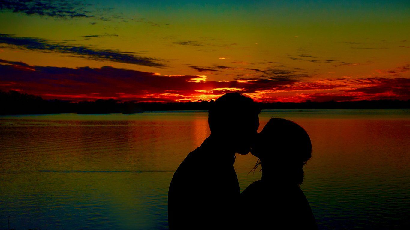 A man and a woman are kissing in front of a sunset over a body of water.