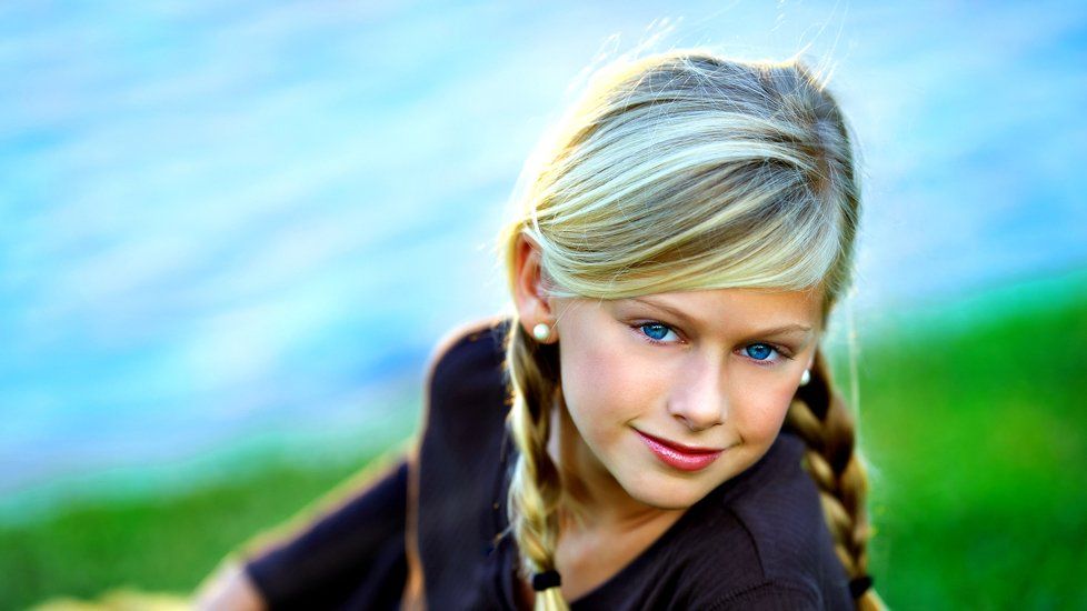 A young girl with braids is sitting in the grass near the water.