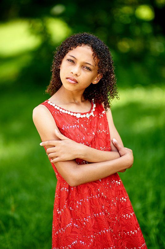 A young girl in a red dress is standing in the grass with her arms crossed.