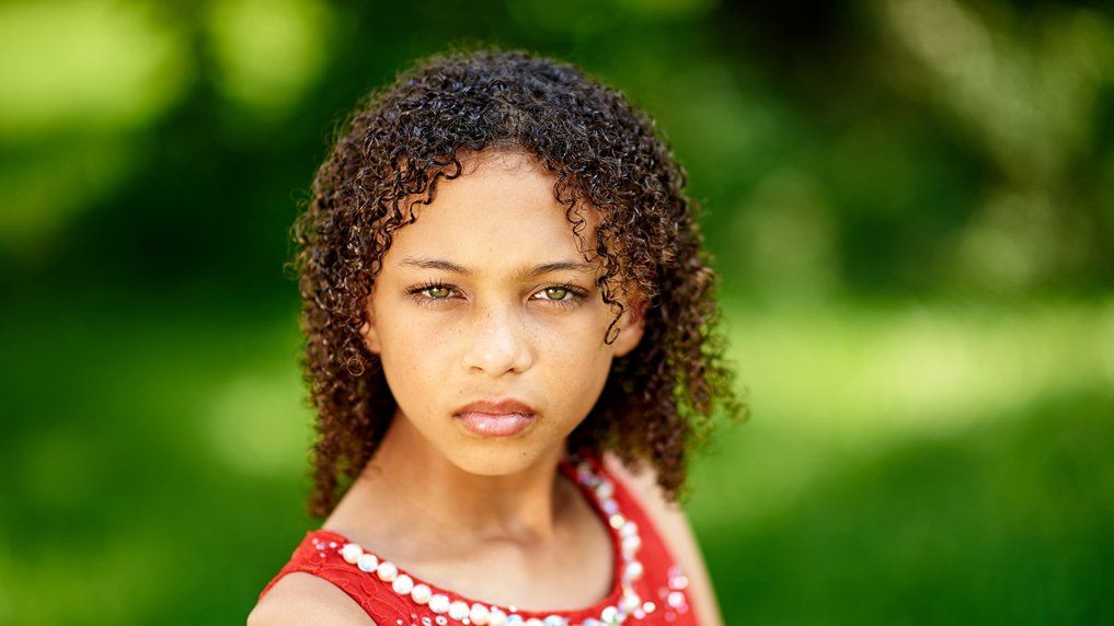A young girl with curly hair is wearing a red dress and looking at the camera.