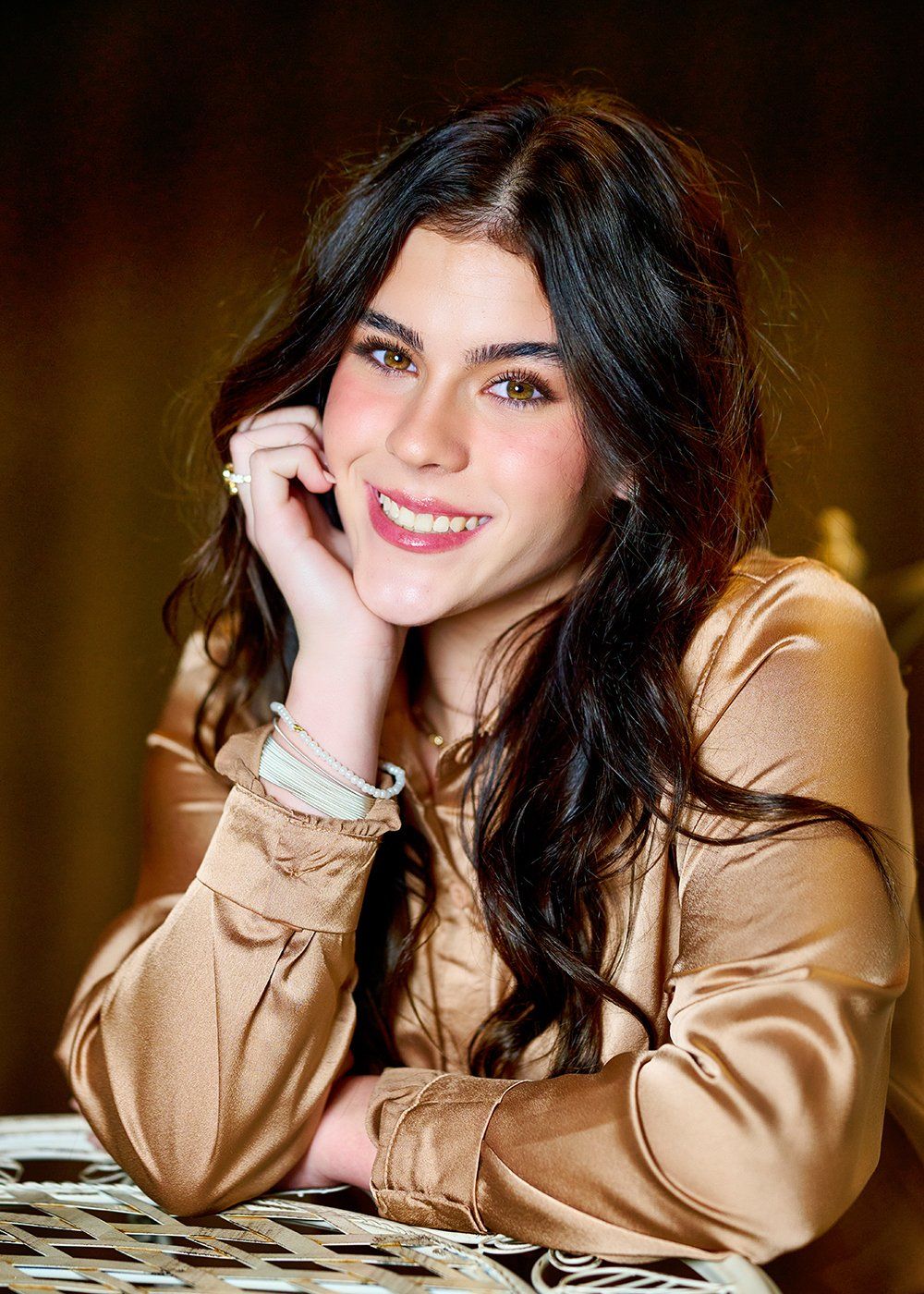 A woman is sitting at a table with her hand on her chin and smiling.