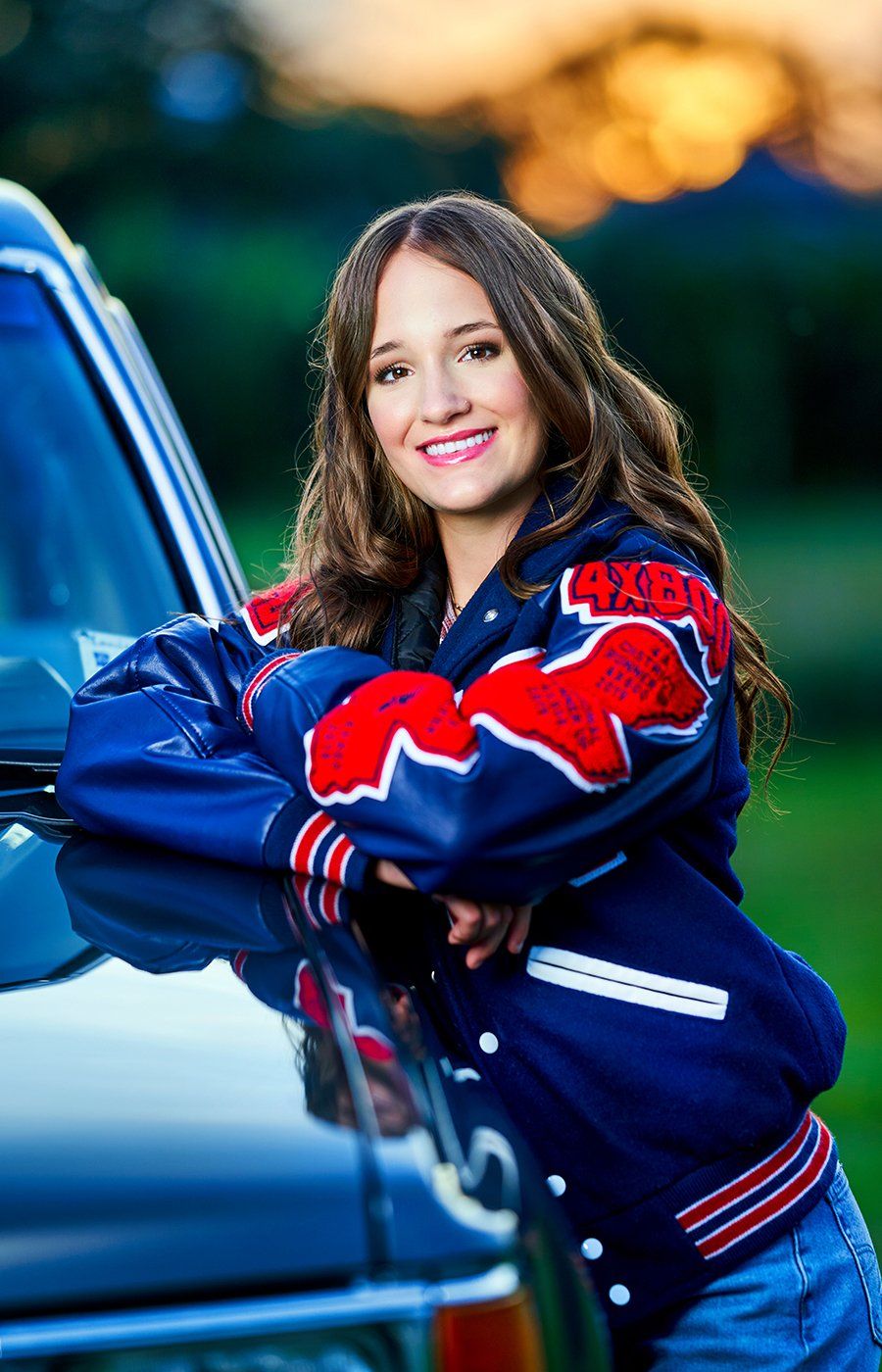 A woman in a blue and red jacket is leaning on the hood of a car.