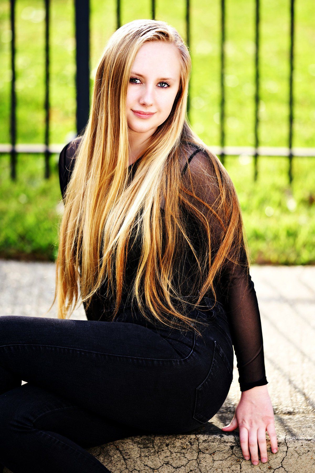 A young woman with long blonde hair is sitting on a rock in front of a fence.