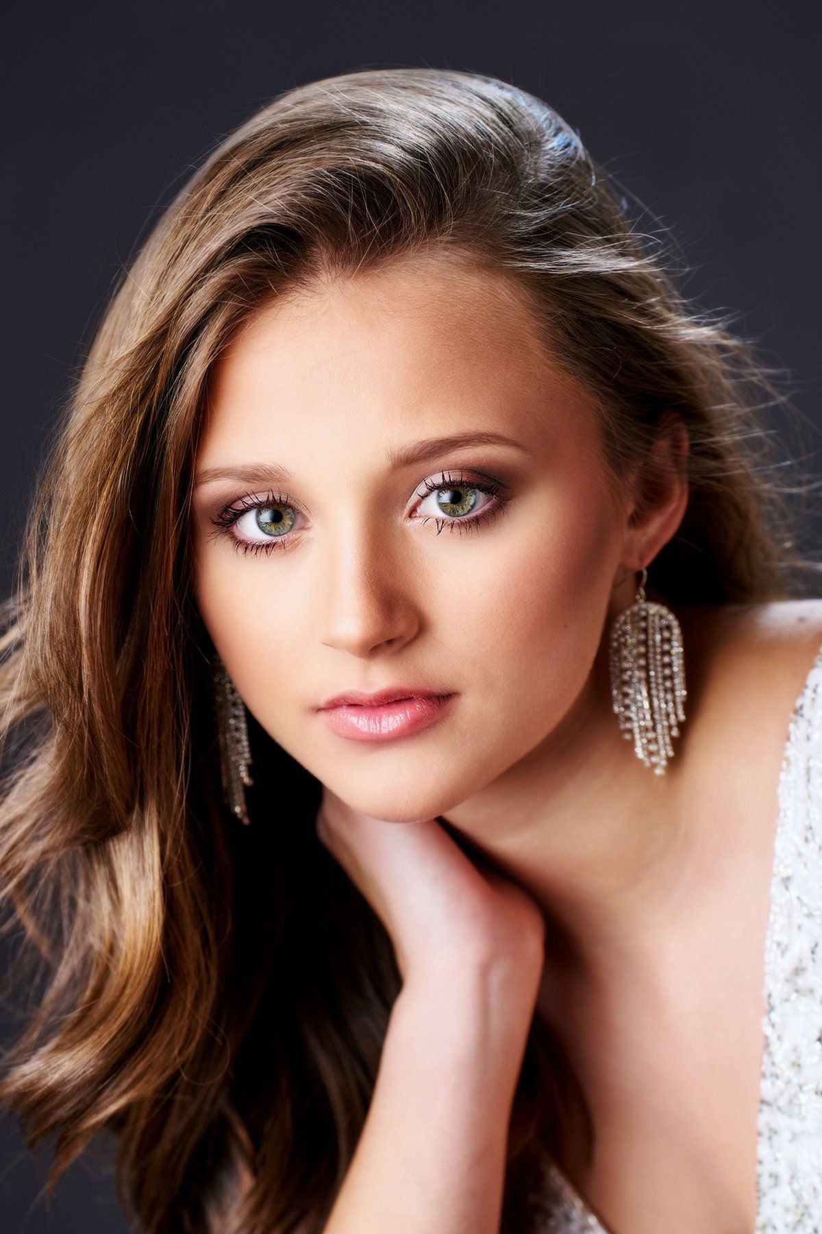 The woman is wearing a white dress and earrings and has her hand on her chin.